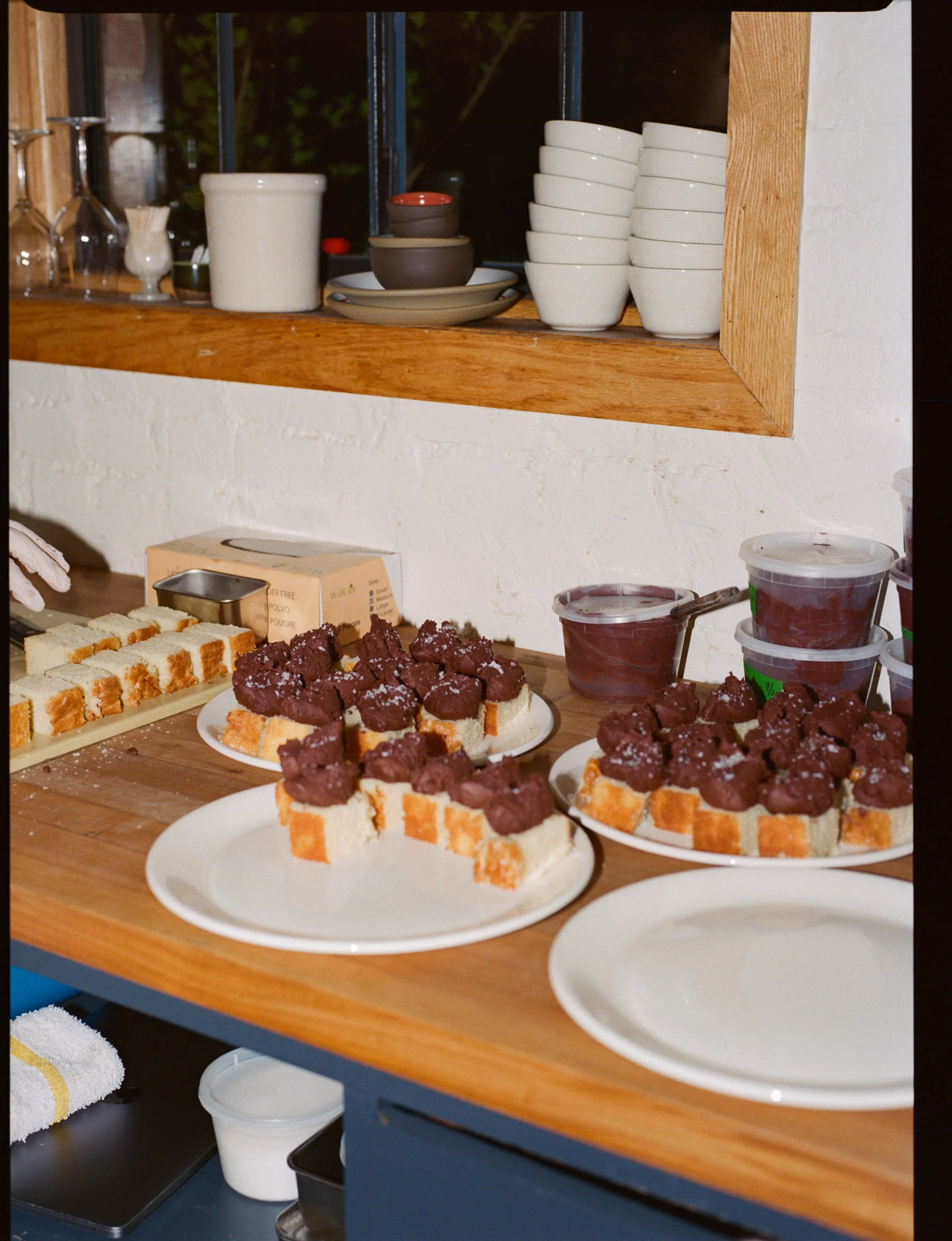 Assorted slices of cake topped with chocolate, displayed on white plates on a wooden table, with bowls of dessert and other kitchen items in the background.