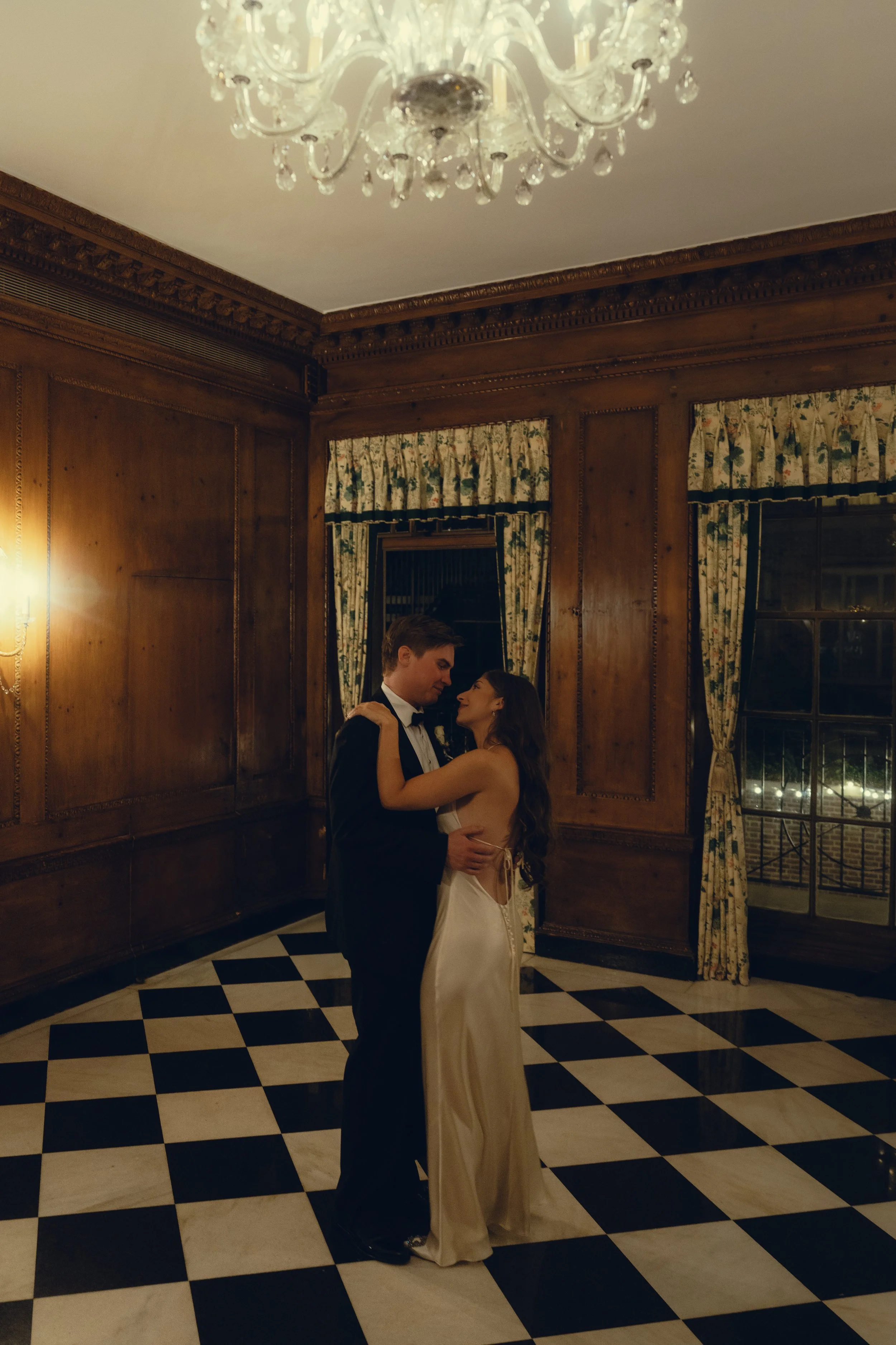 A couple dancing in formal attire in a wood-paneled room with checkered black and white floor, floral curtains, and a chandelier.
