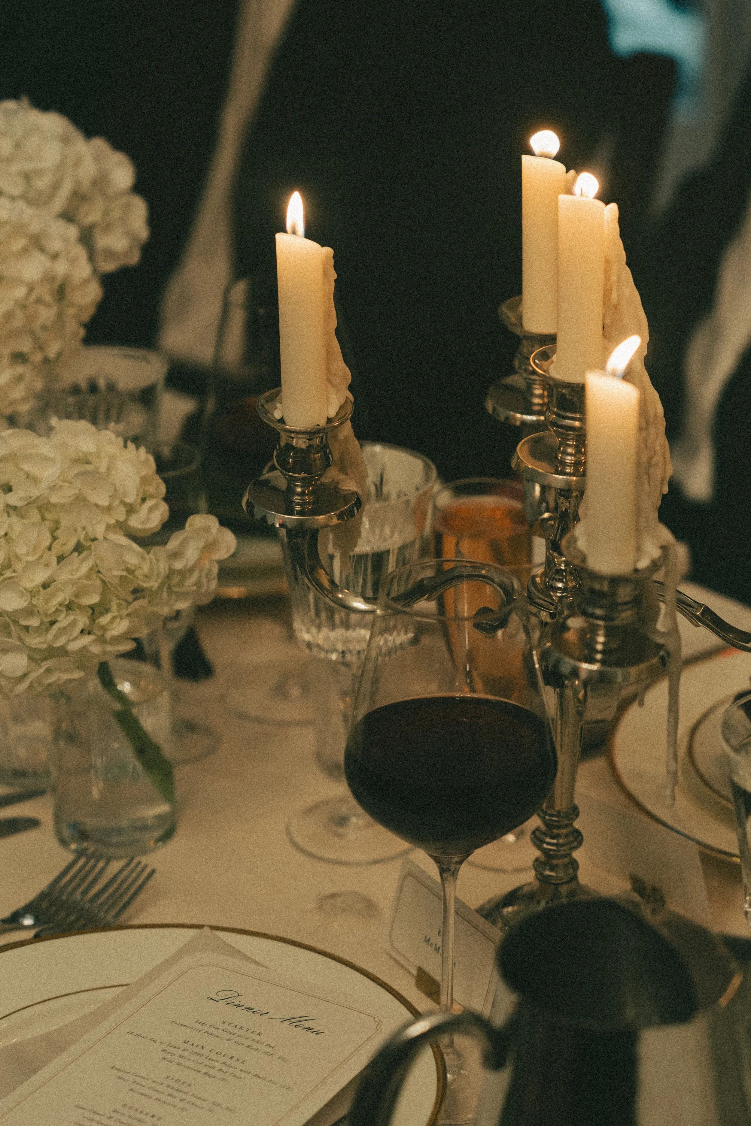 A dinner table with a glass of red wine, a candelabrum with lit white candles, flowers, and a menu.