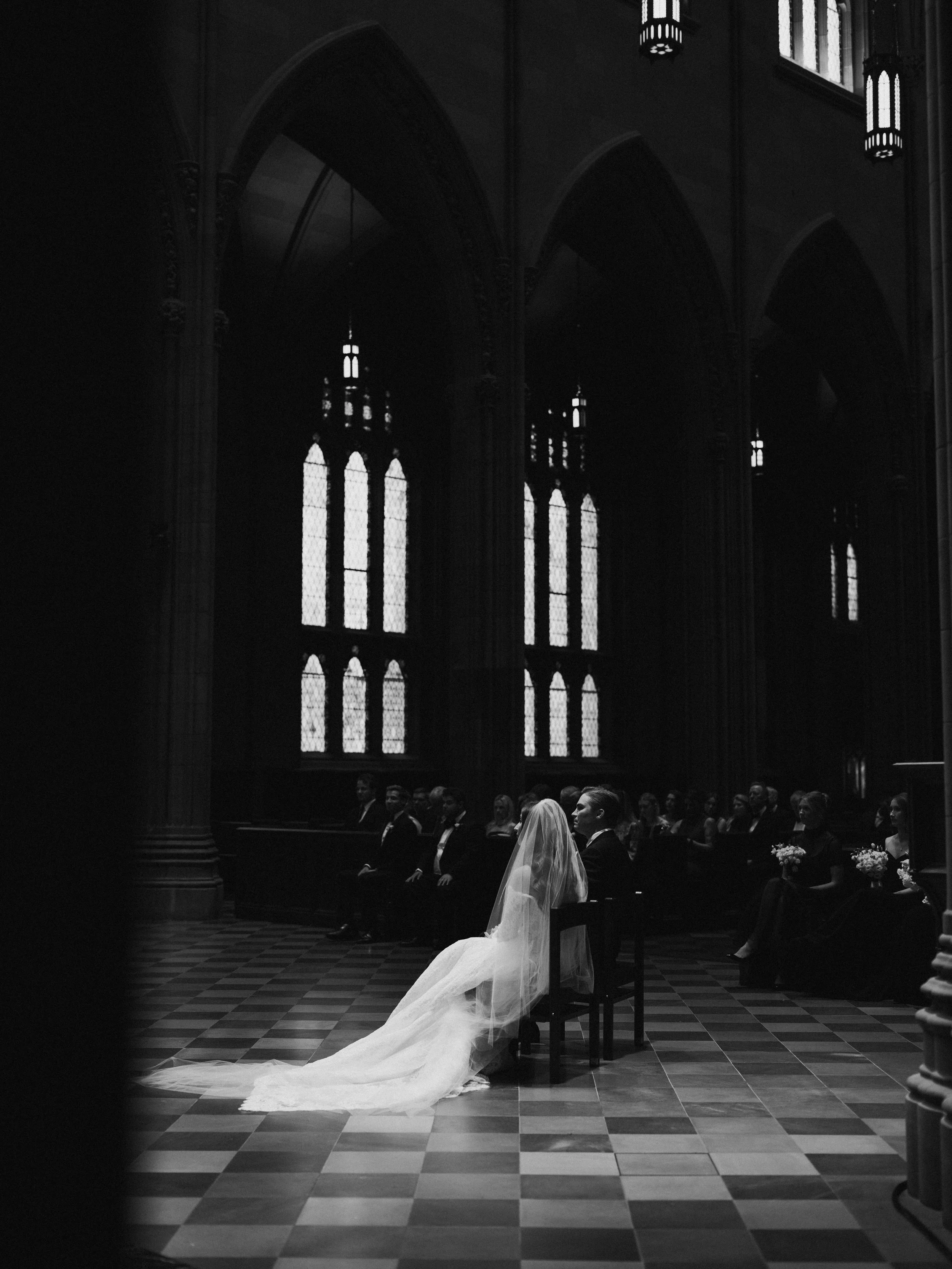A bride in a wedding dress with a veil sitting on a chair in a church during a wedding ceremony, with guests seated behind her. The church has gothic architecture with high arches and stained glass windows.