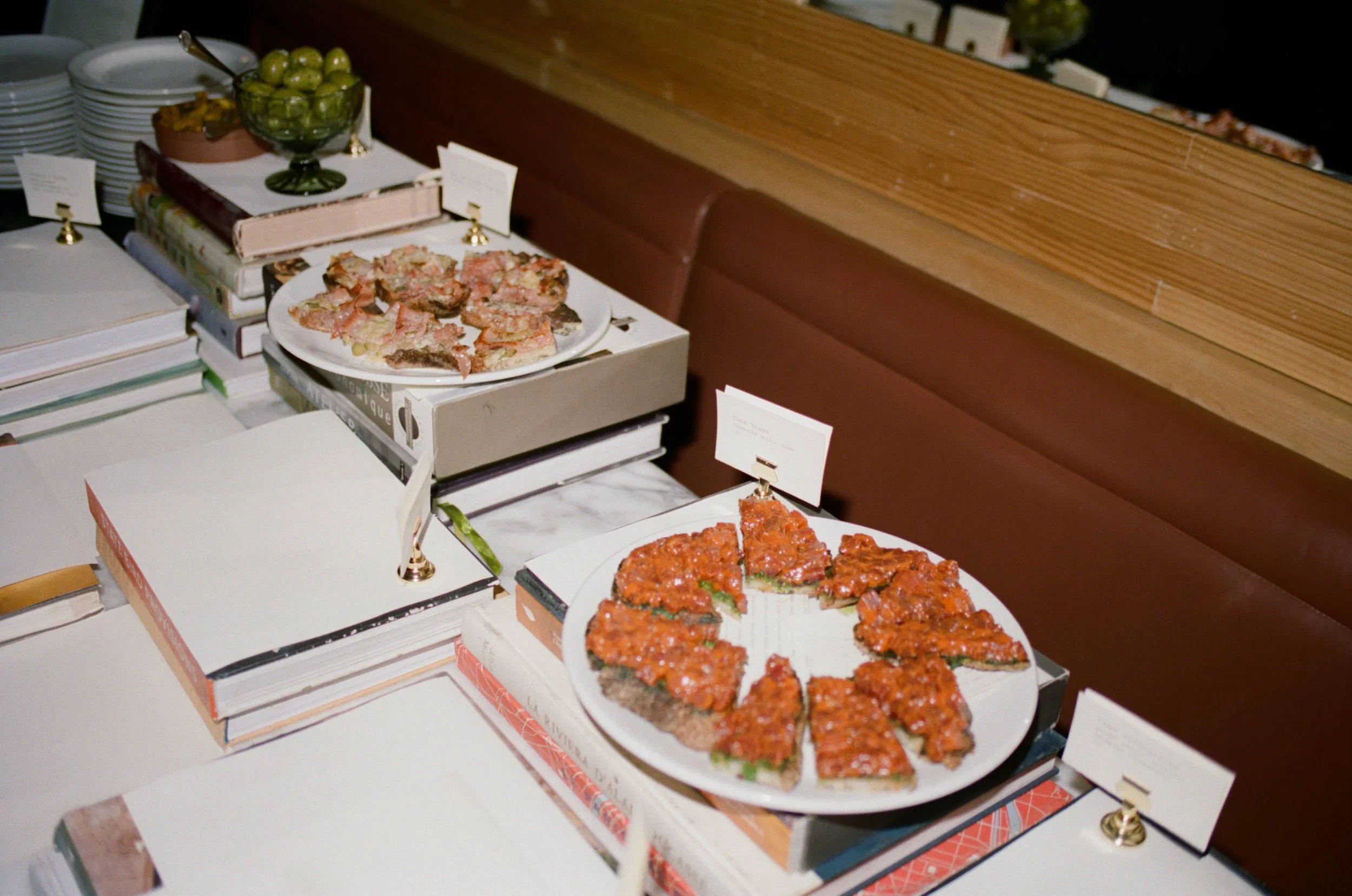 A table display of various food items, including a plate of baked eggplant slices topped with tomato sauce, a bowl of green grapes, and a plate of sliced bread, with stacks of books and place cards surrounding the dishes.