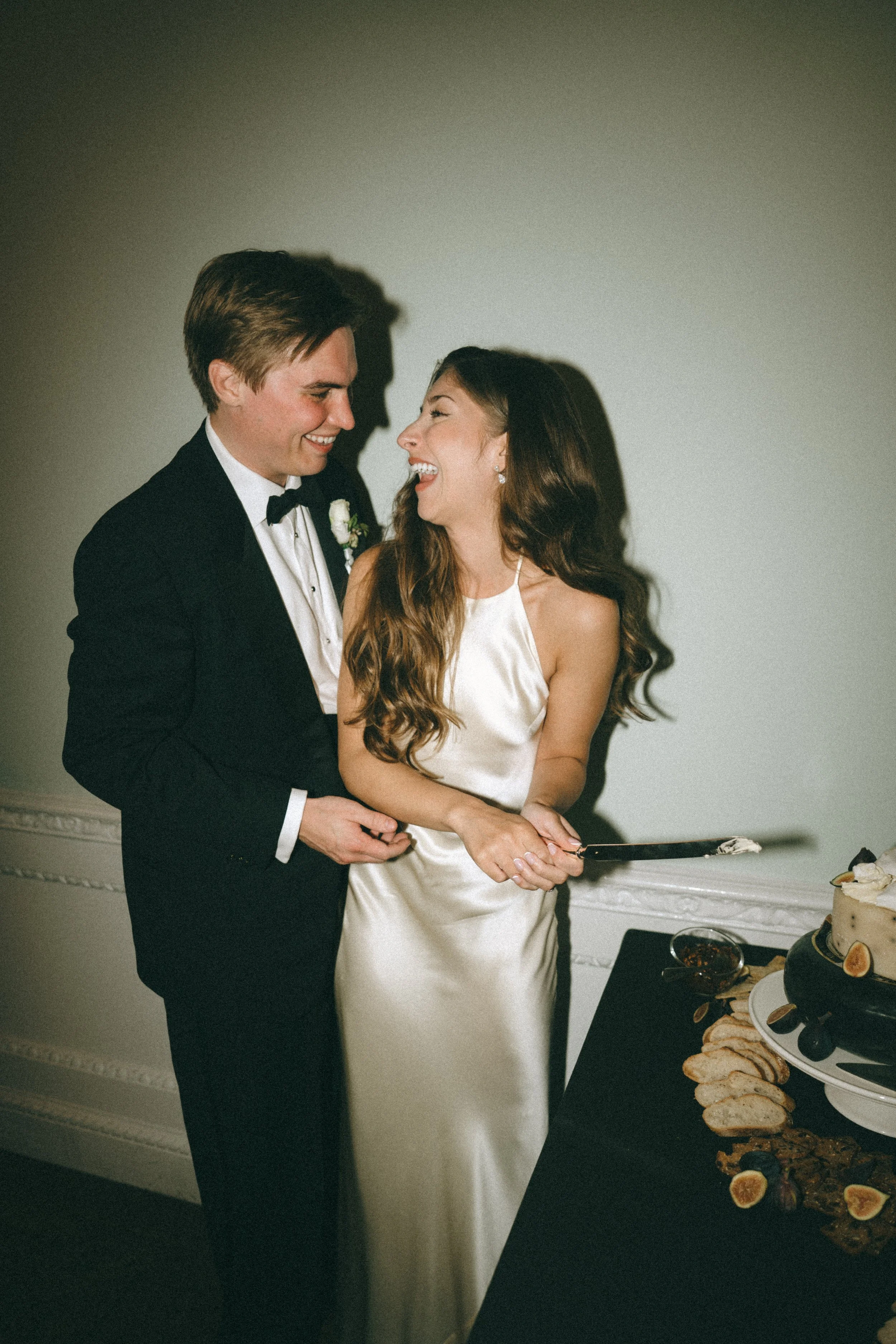 A bride and groom in wedding attire laughing and smiling as they cut their wedding cake together.