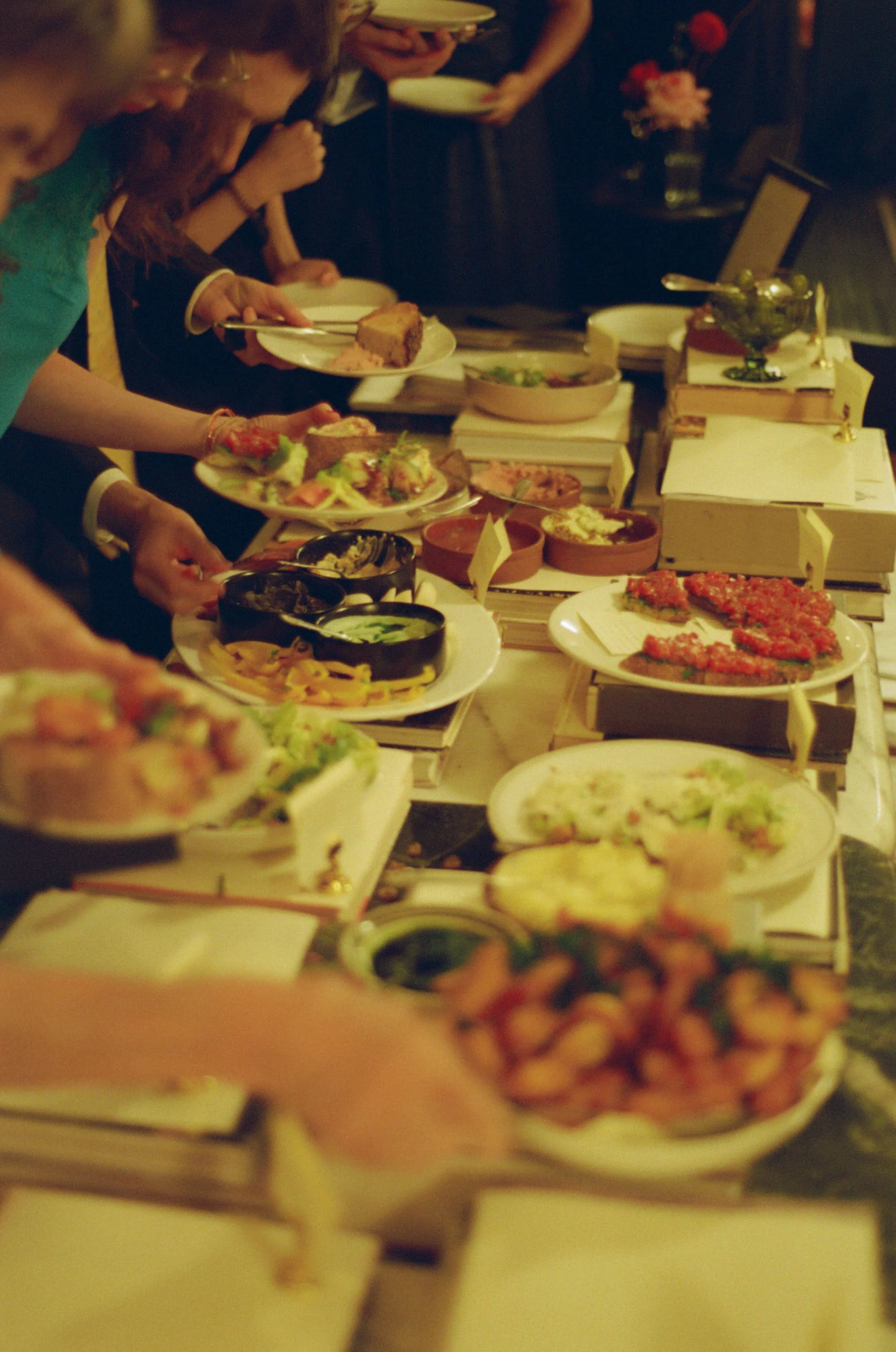 People at a buffet table selecting various foods, with plates and bowls of dishes including salads, desserts, and appetizers.
