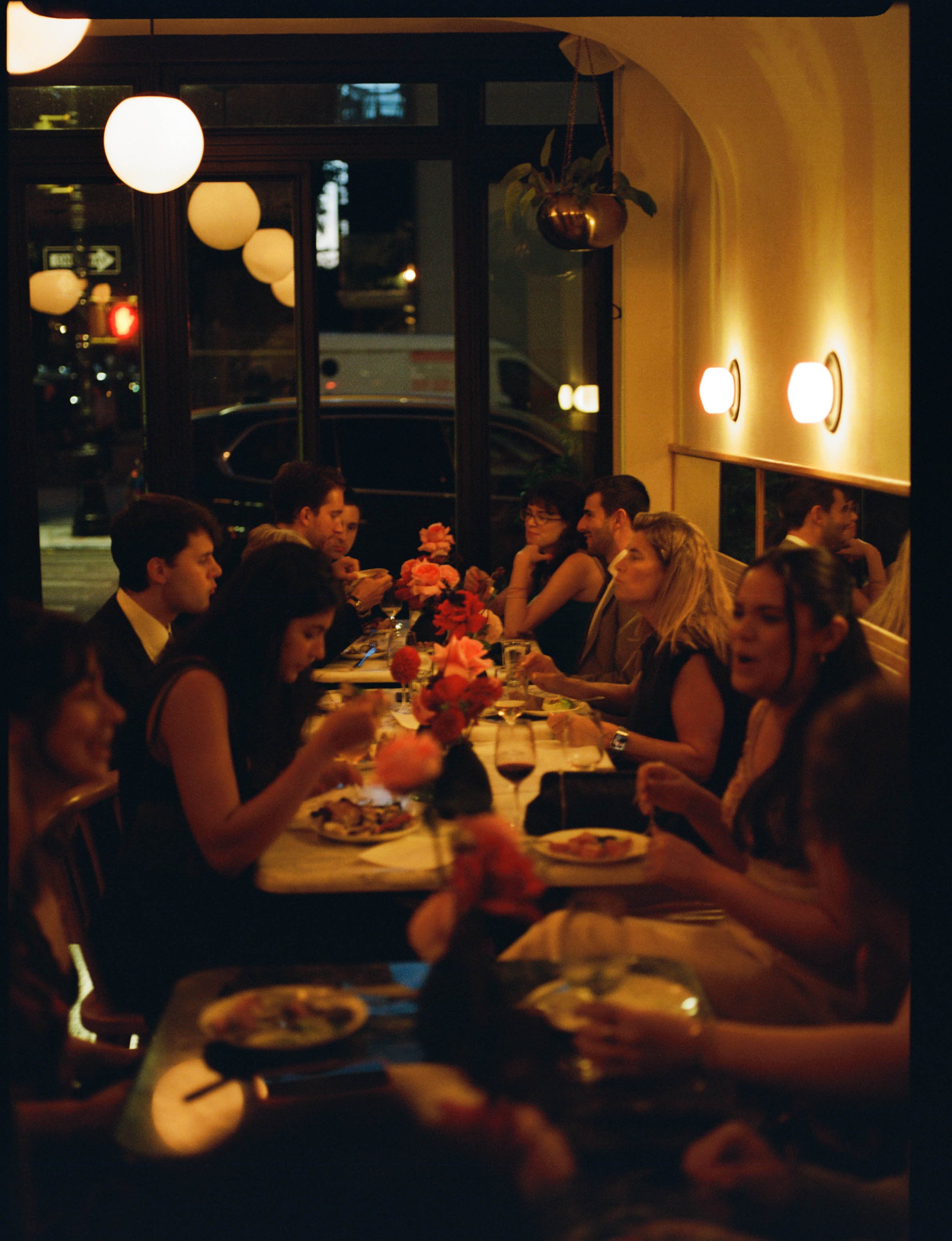 People dining at a restaurant table during the evening, with floral centerpieces and glasses of wine, lit by soft warm lighting.