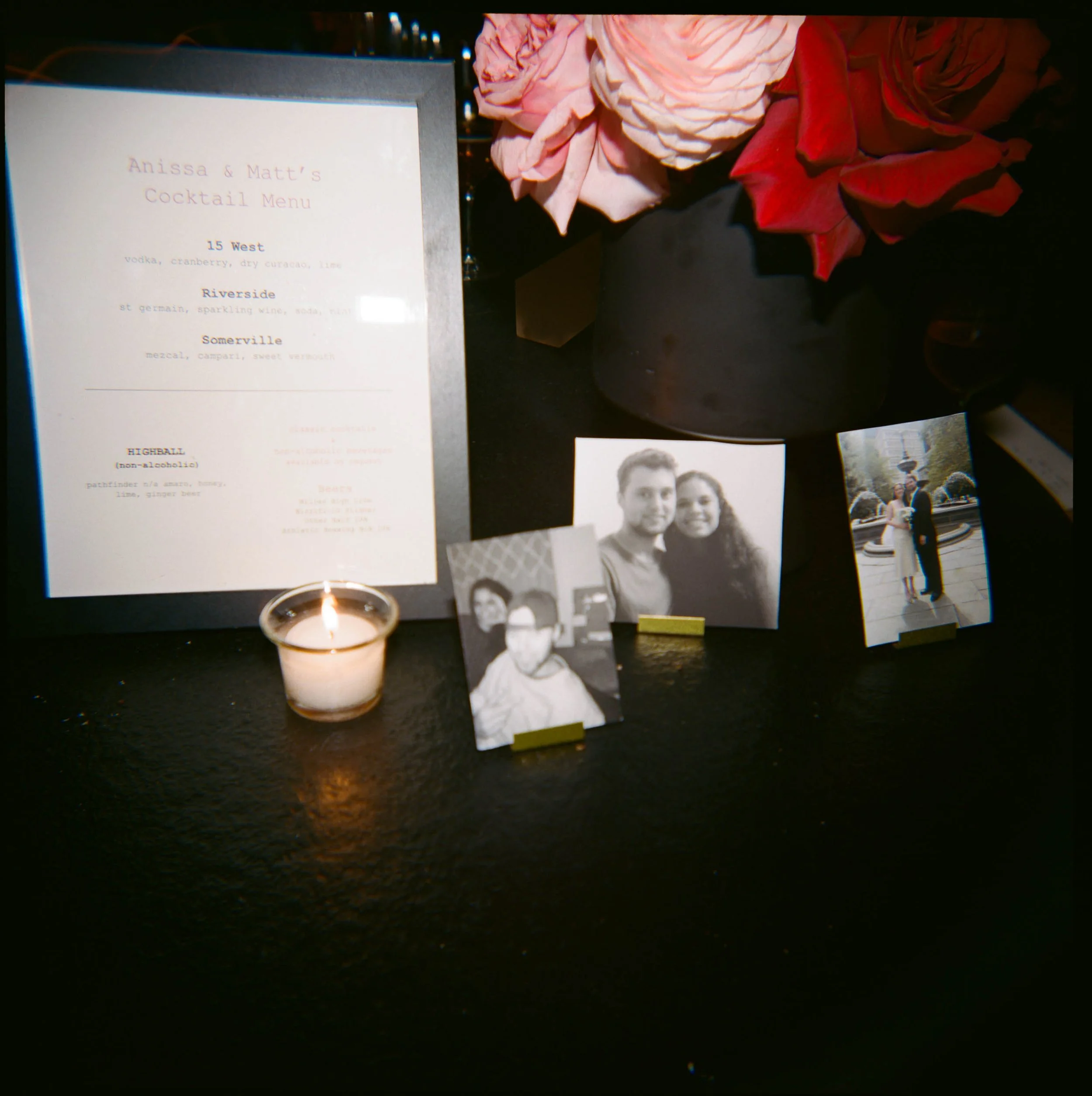 A memorial display includes a cocktail menu, a lit candle, a black vase with pink and red flowers, and several black-and-white photographs of people.