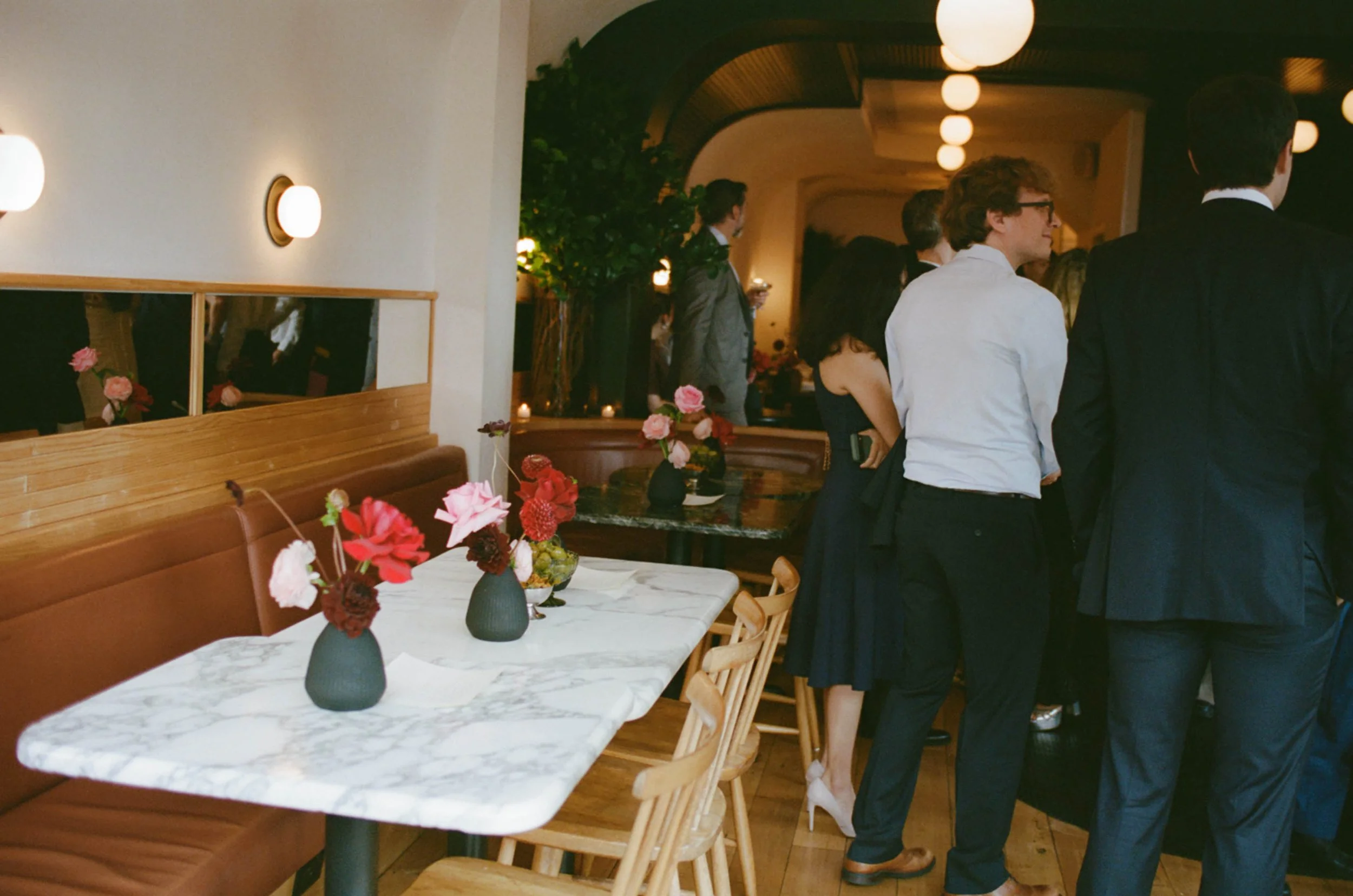 People dressed in formal attire gather at a restaurant or event space with a marble table, floral arrangements, and ambient lighting.