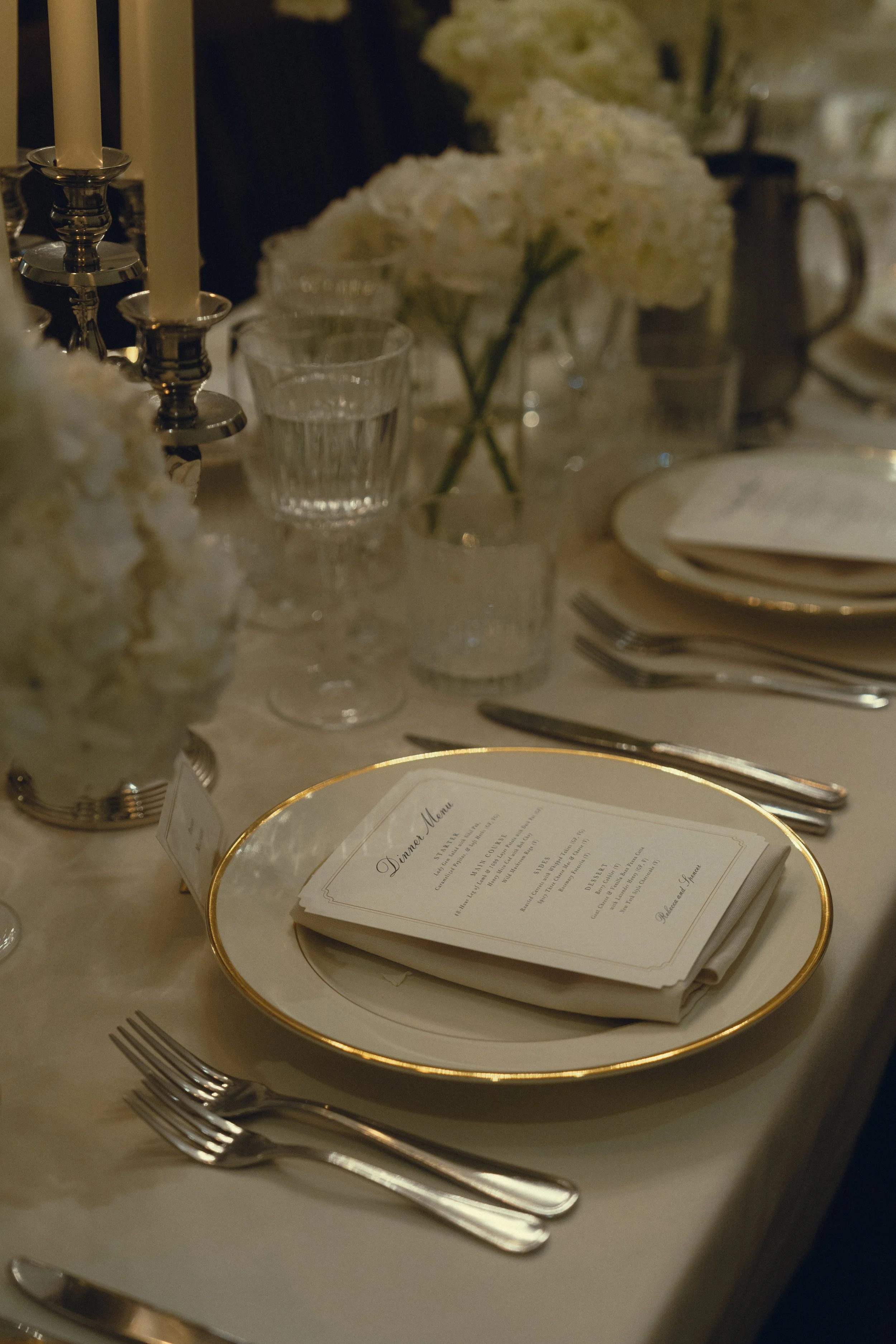 Elegant dining table setting with a white plate with gold trim, folded napkin with a printed menu on top, silverware, drinking glasses, white floral centerpiece, and tall white candles in silver candlesticks.