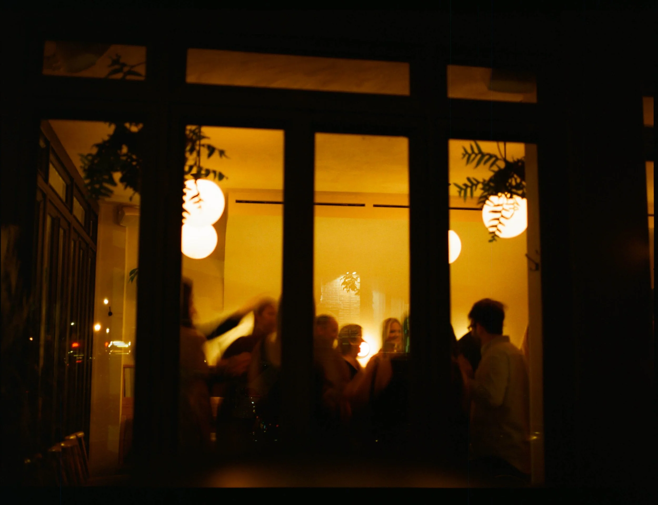 People socializing inside a warmly lit restaurant or cafe seen through a window at night.