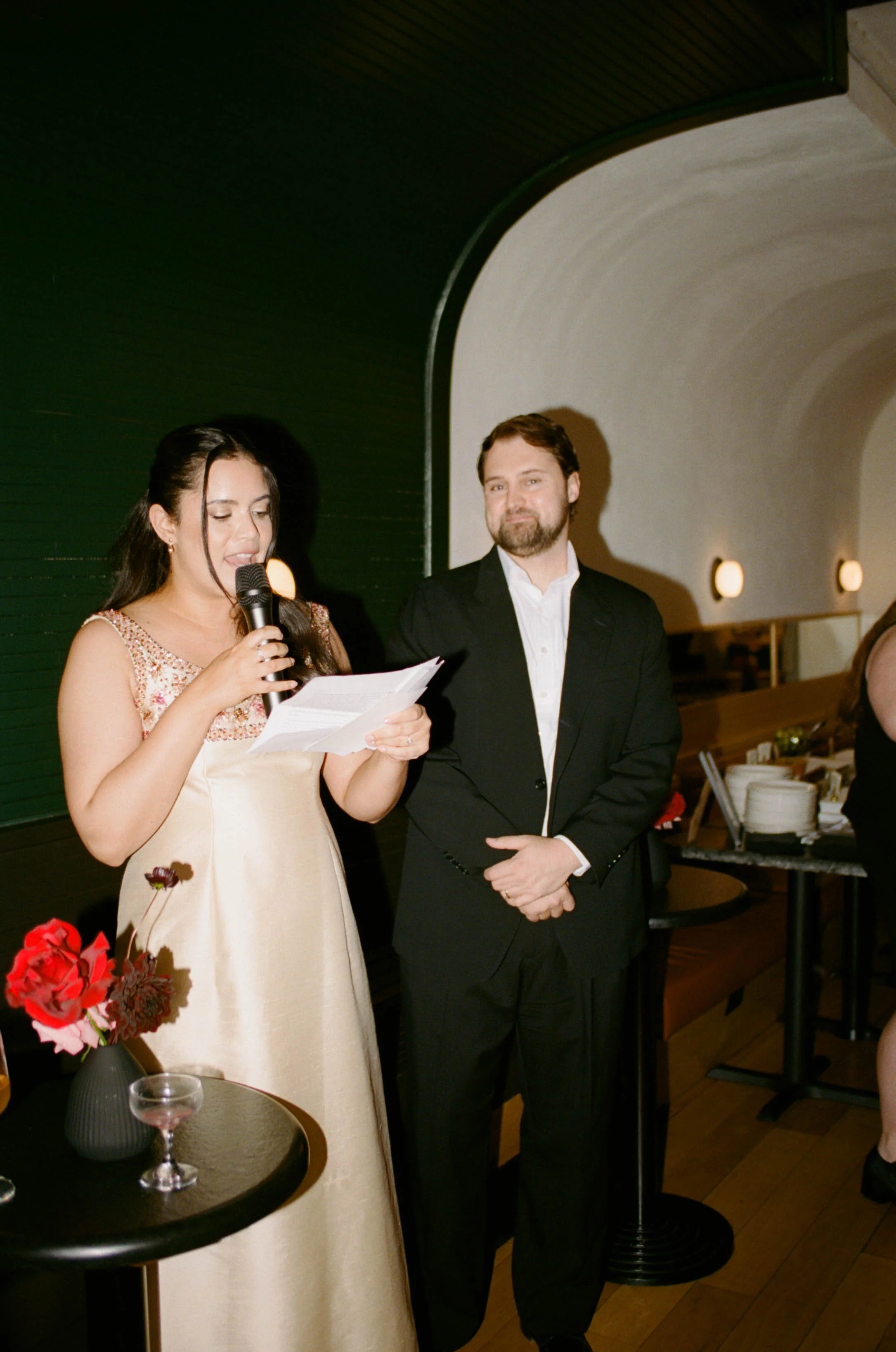 A woman in a champagne-colored dress reading from a paper while holding a microphone, standing next to a man in a black suit and white shirt at an indoor event.