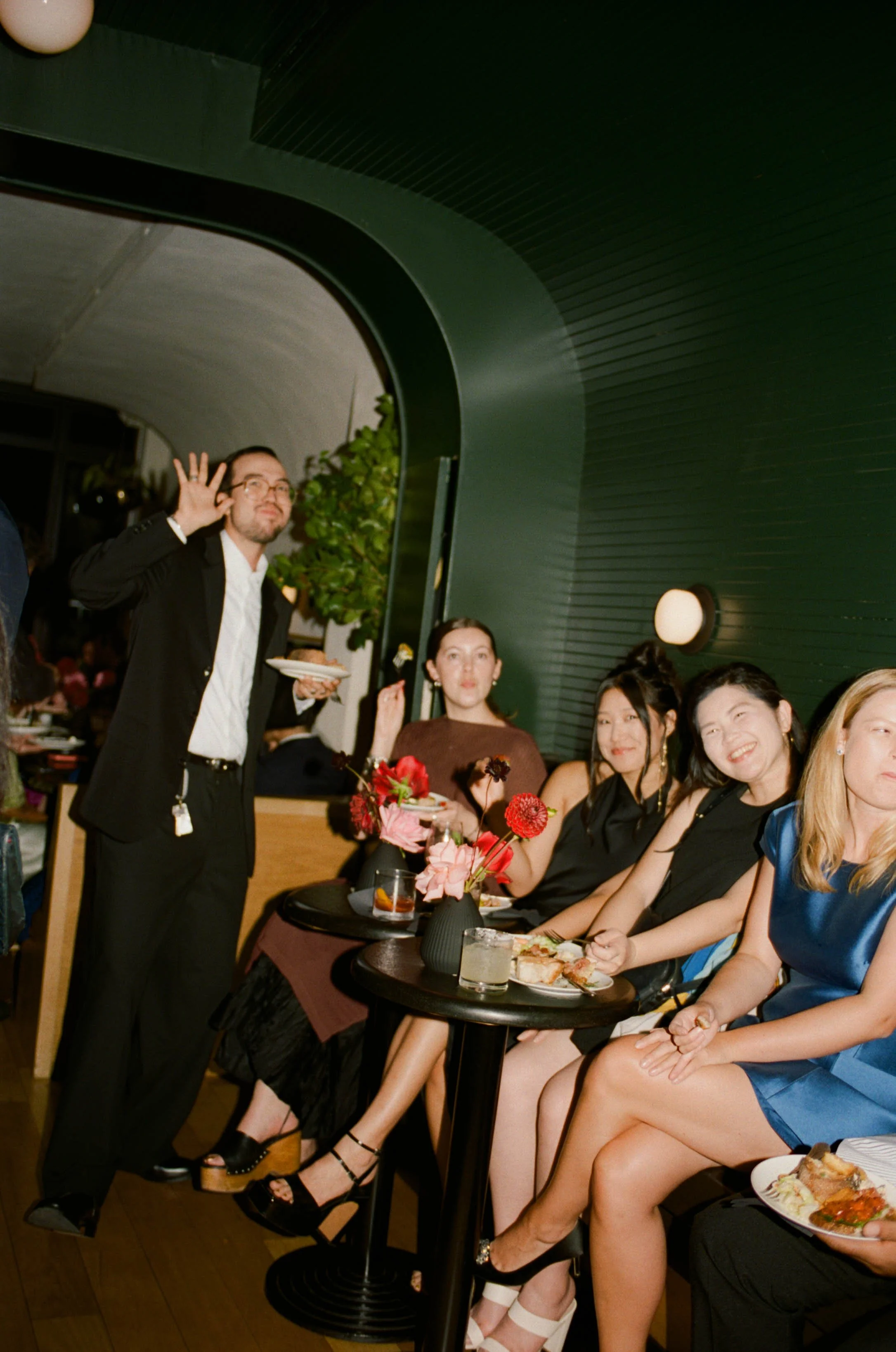 People sitting at a table with food and drinks, celebrating at a party in a modern venue with dim lighting and green walls.