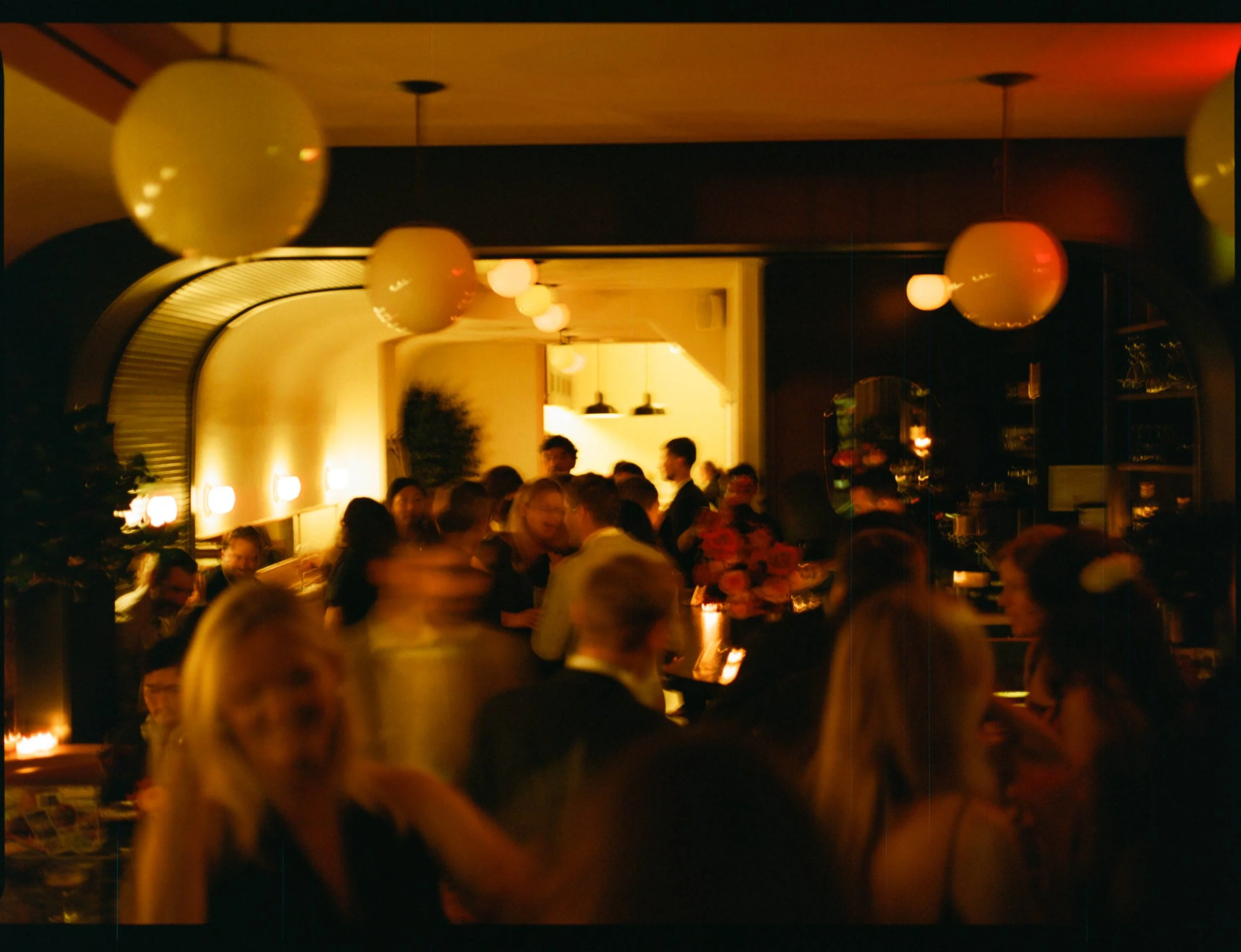 A crowded indoor party at night with warm lighting, people socializing, and hanging yellow balloons.