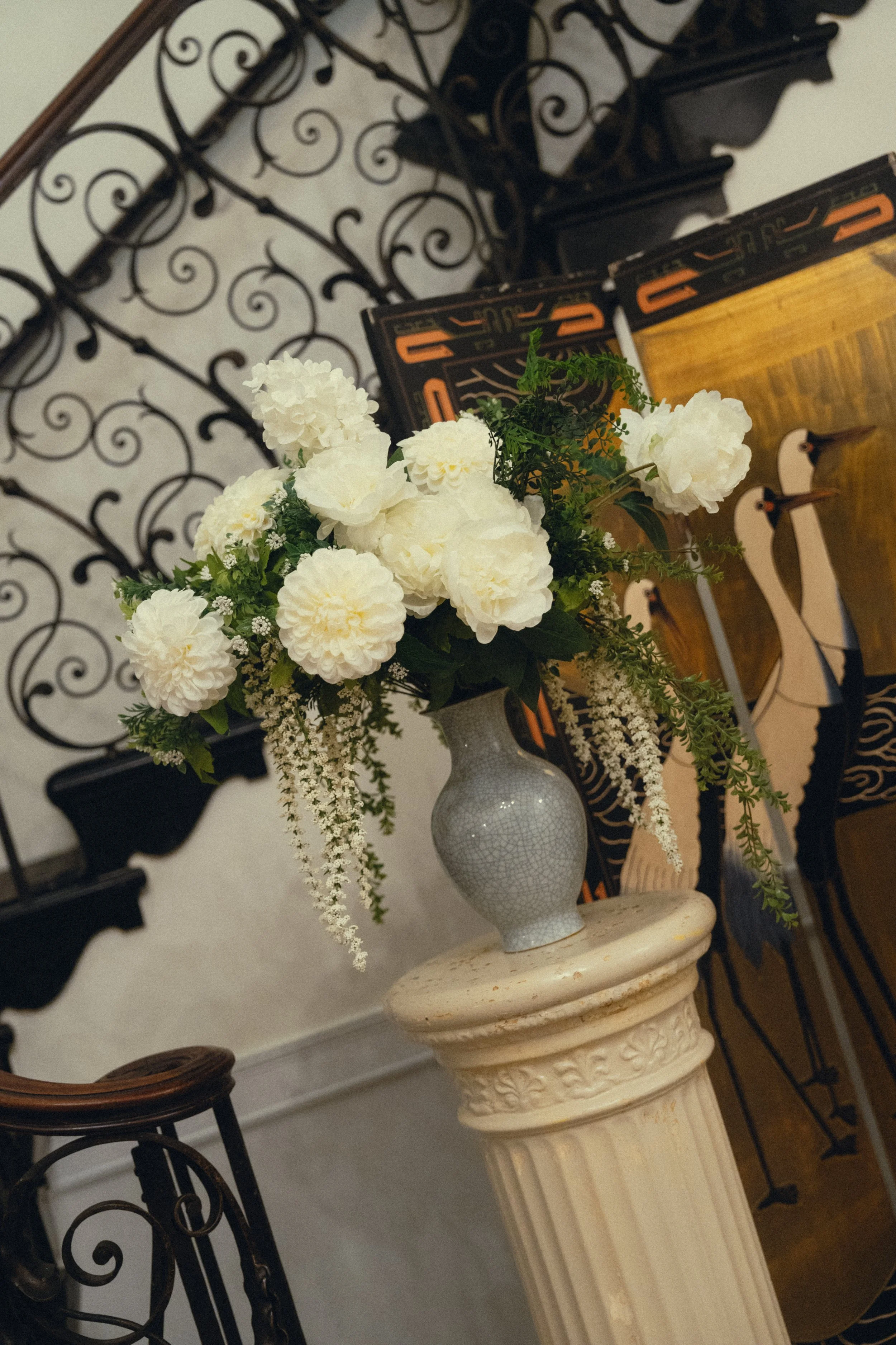 A white ceramic vase with crackle glaze holding a bouquet of white flowers, including roses and other blooms, on a cream-colored pedestal with decorative carvings, placed in front of a wooden and wrought-iron decorative wall art and a painting of sty