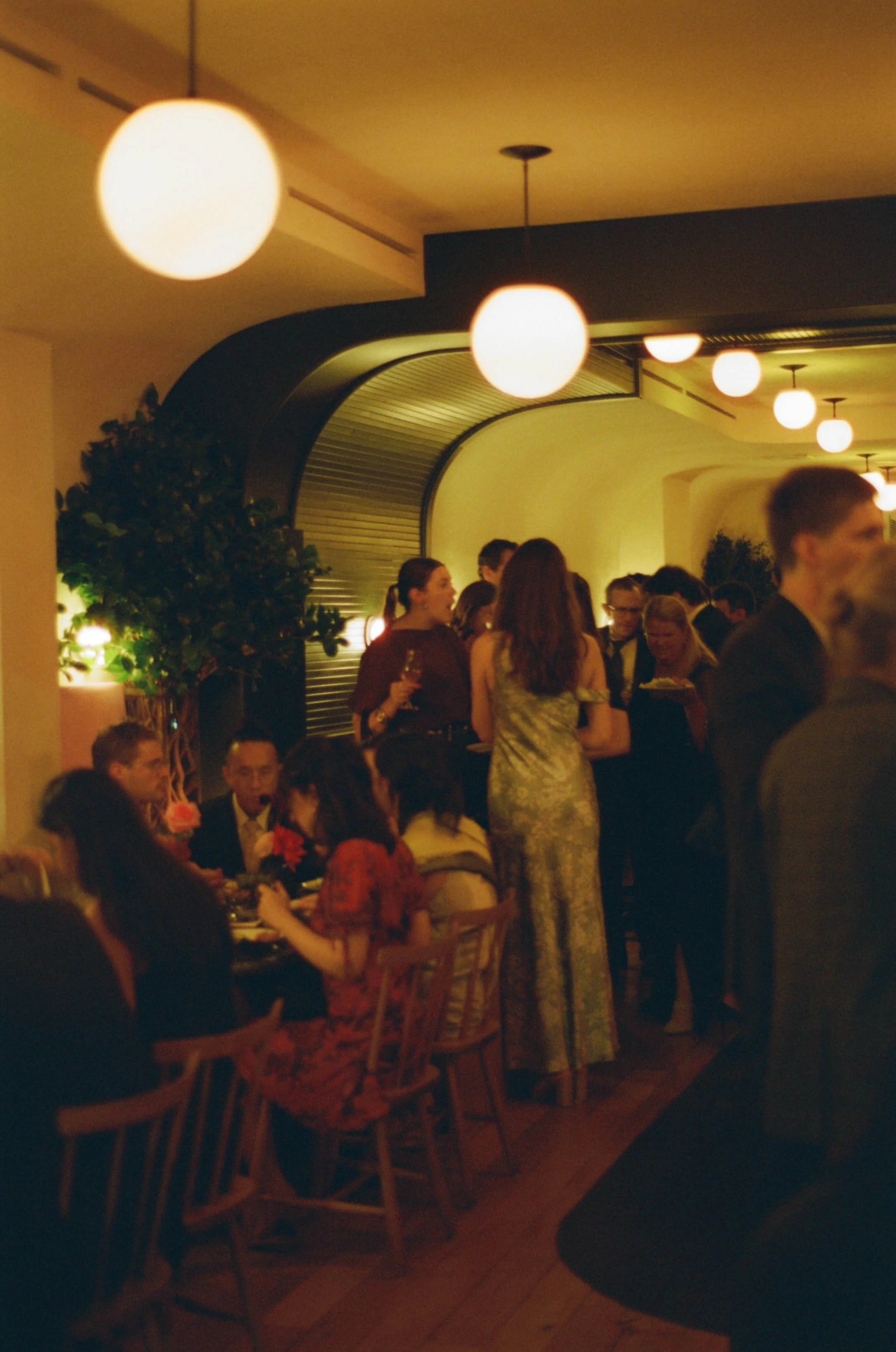 A social gathering in an elegant indoor setting with people standing and sitting, some holding drinks and plates, under soft lighting with round ceiling lights.