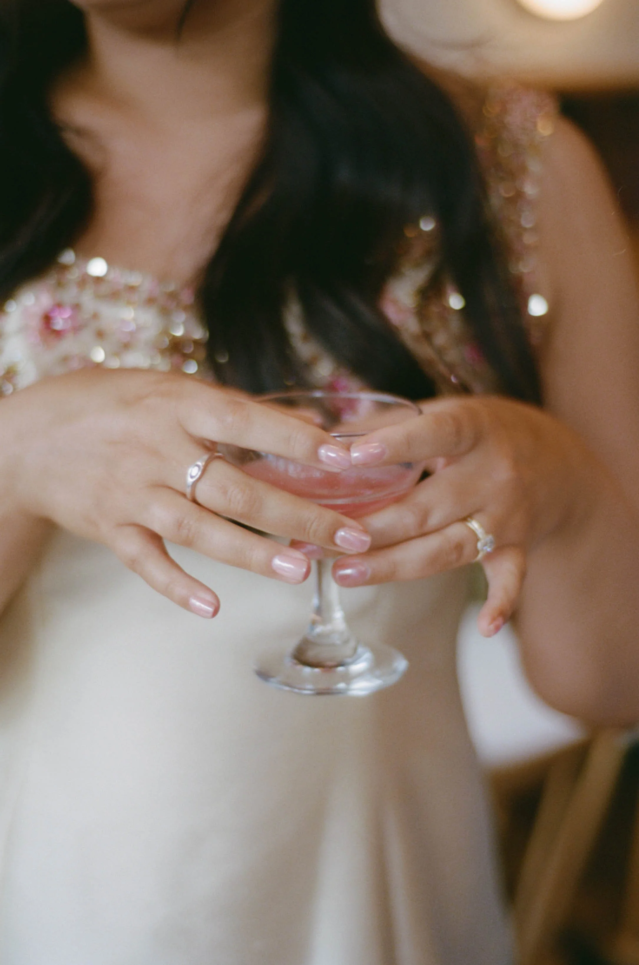 Person holding a pink-colored cocktail in a glass with both hands, wearing rings, and dressed in a sparkly outfit.