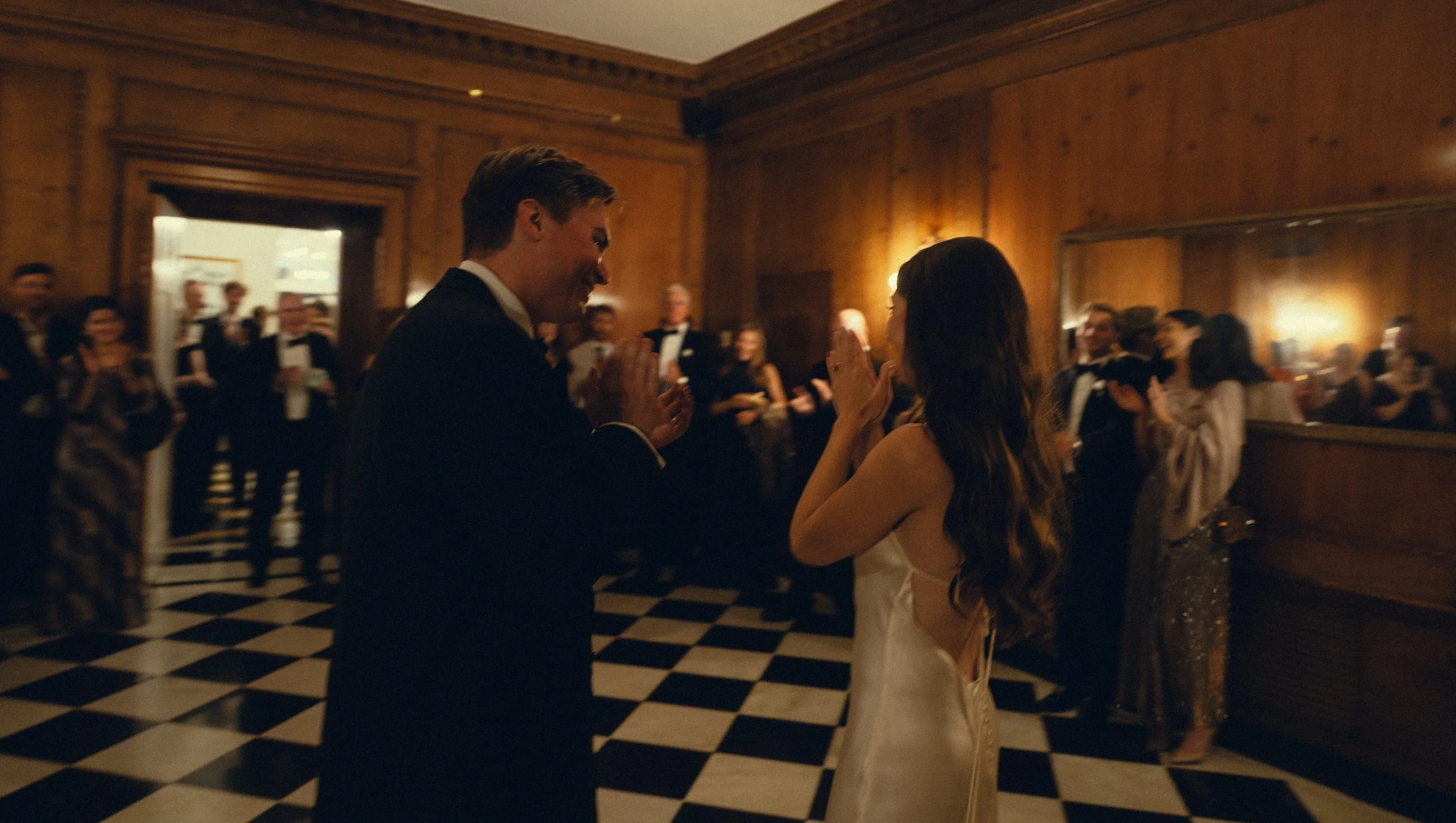 A man and woman dancing at a formal event, surrounded by guests in tuxedos and dresses, in a wood-paneled room with a checkered black-and-white floor.
