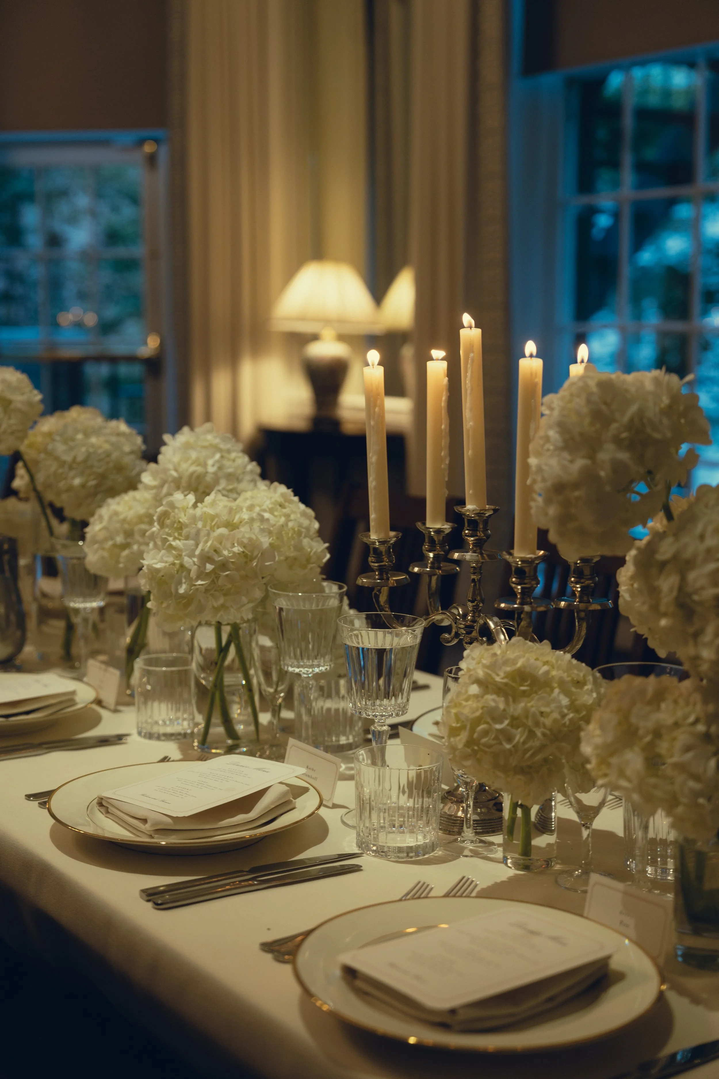 Elegant dining table set with white floral centerpieces, tall white candles in silver candlesticks, wine glasses, and menus, with warm lighting and a window with curtains in the background.