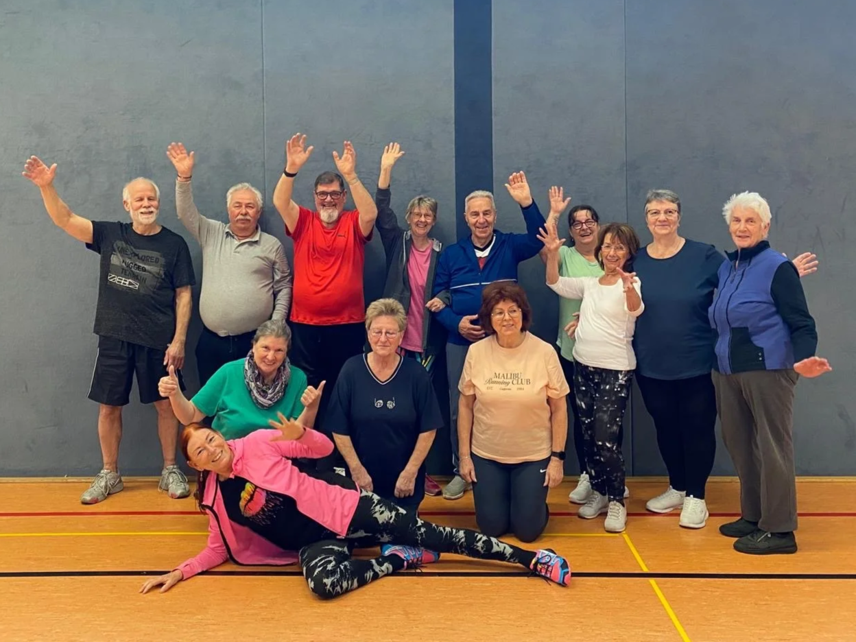 Gruppe älterer Menschen in Sportkleidung in einer Turnhalle stehen zusammen, alle lächeln und winken.