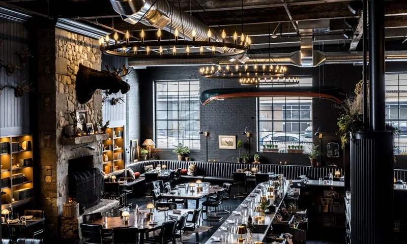 Interior of a modern restaurant with exposed brick walls, large windows, and a stone fireplace topped with a mounted animal head. The space is decorated with chandeliers, candles on tables, and various plants, creating a cozy and stylish ambiance.
