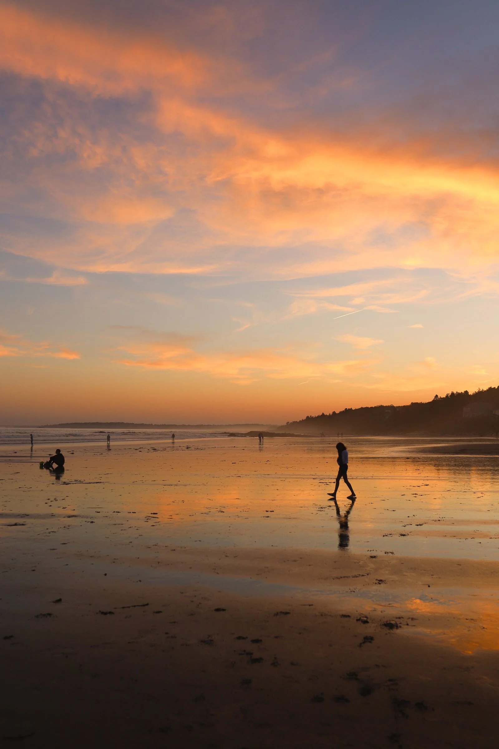 People walking and taking photos on a beach at sunset, with colorful clouds and reflections on wet sand.