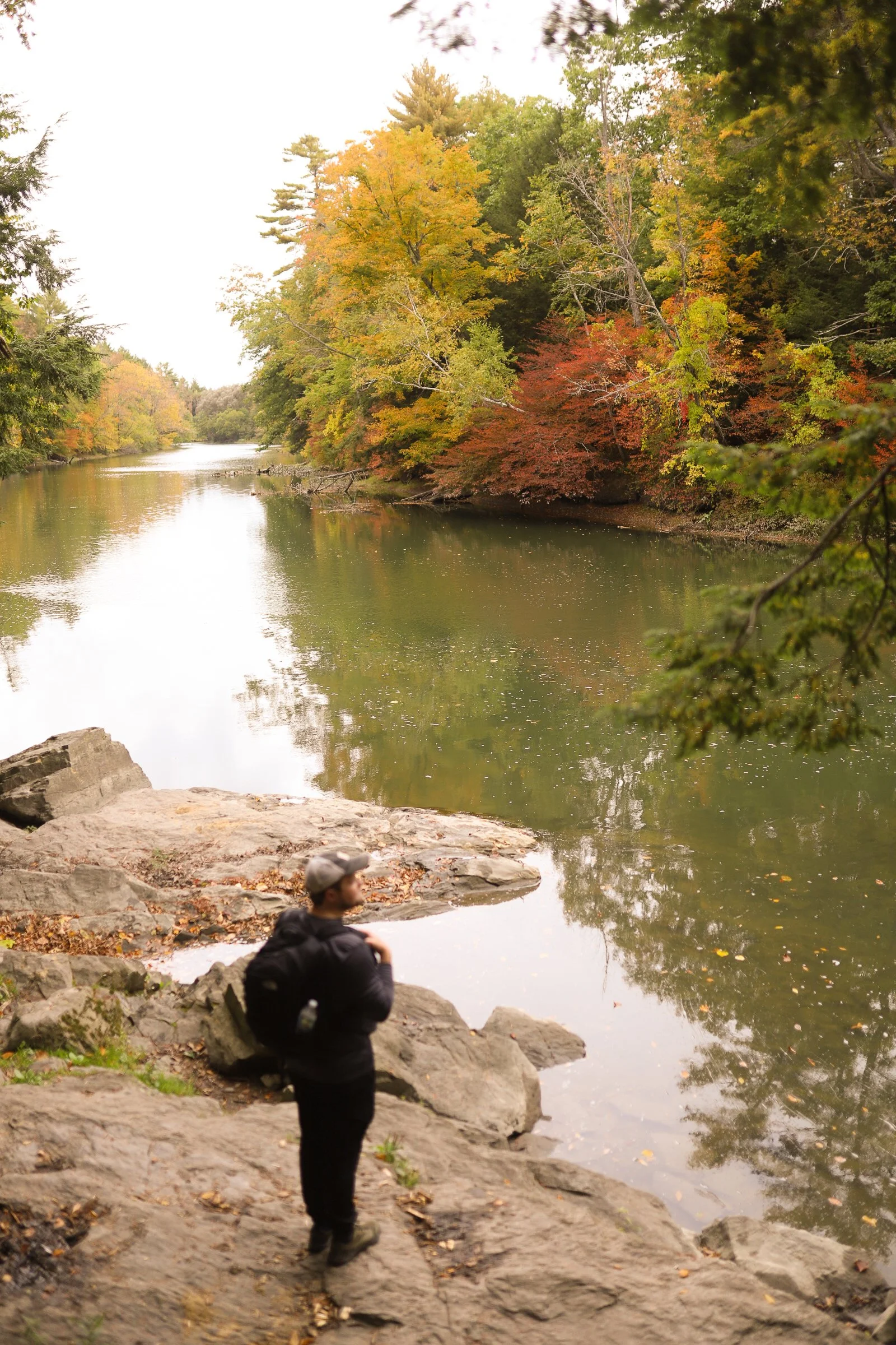 A man standing on rocks by a river surrounded by trees with autumn foliage.