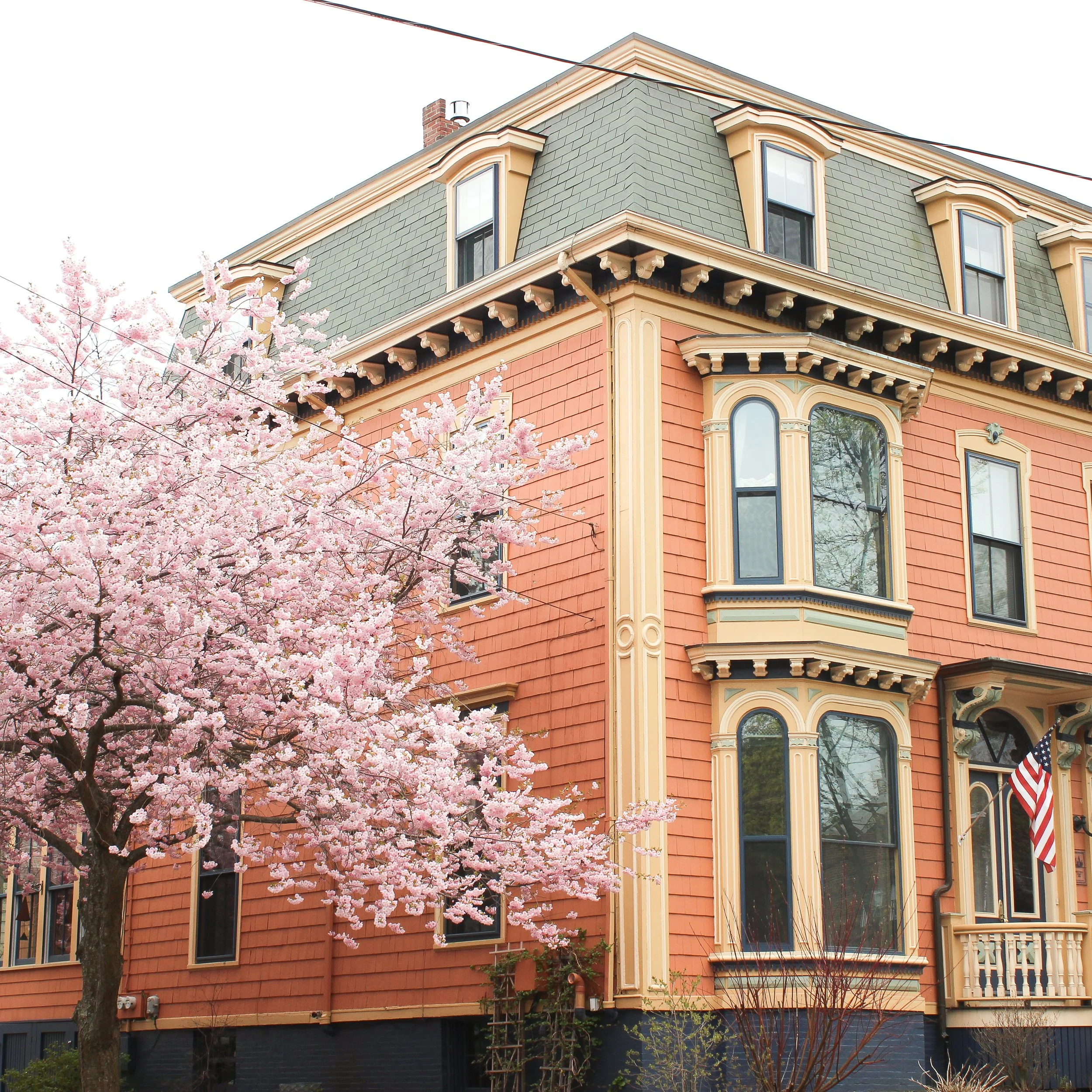 A colorful Victorian-style house with decorative trim, large bay windows, and an American flag, with a blooming pink cherry blossom tree in front.