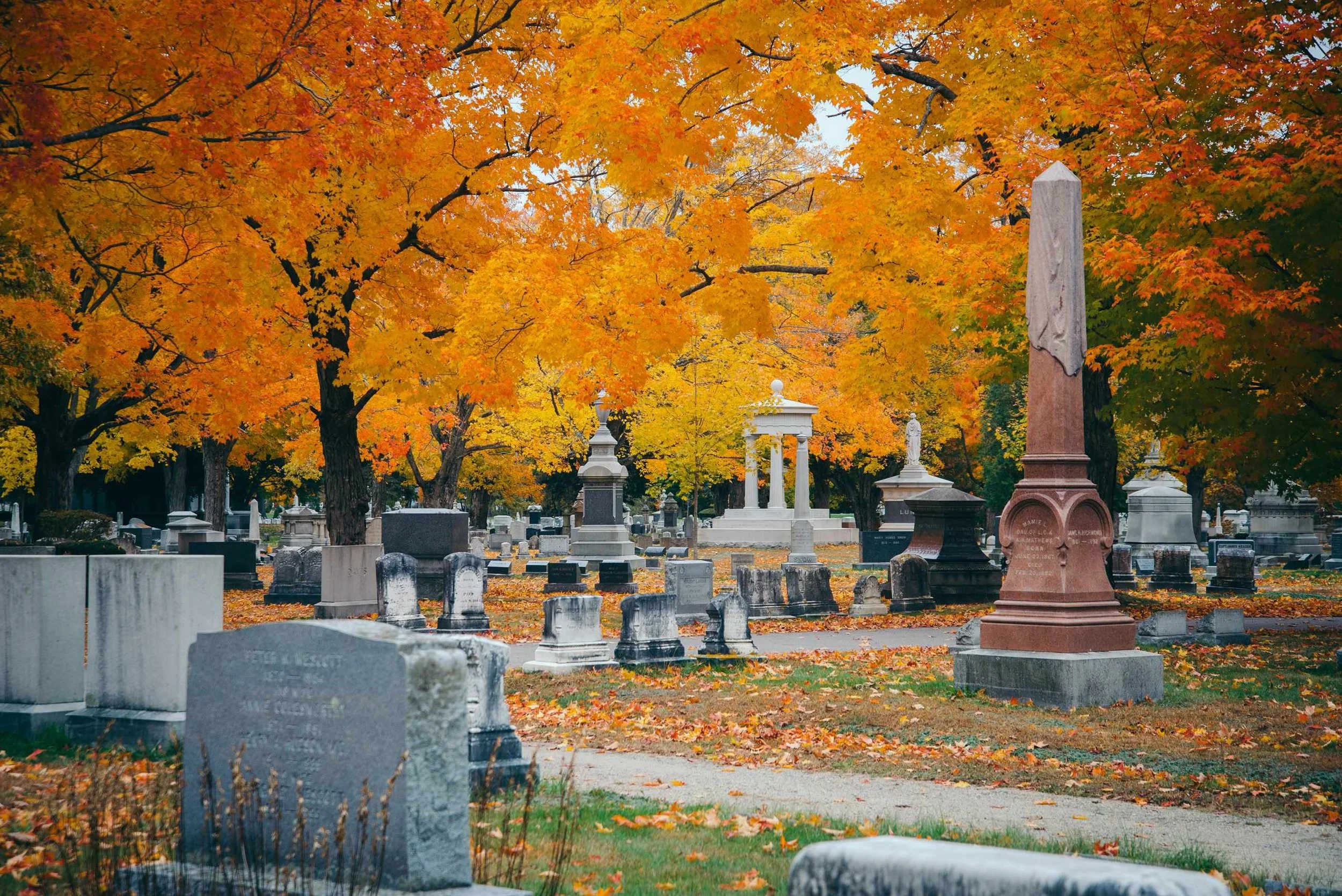 A cemetery with gravestones and monuments surrounded by trees with orange, yellow, and red autumn leaves.