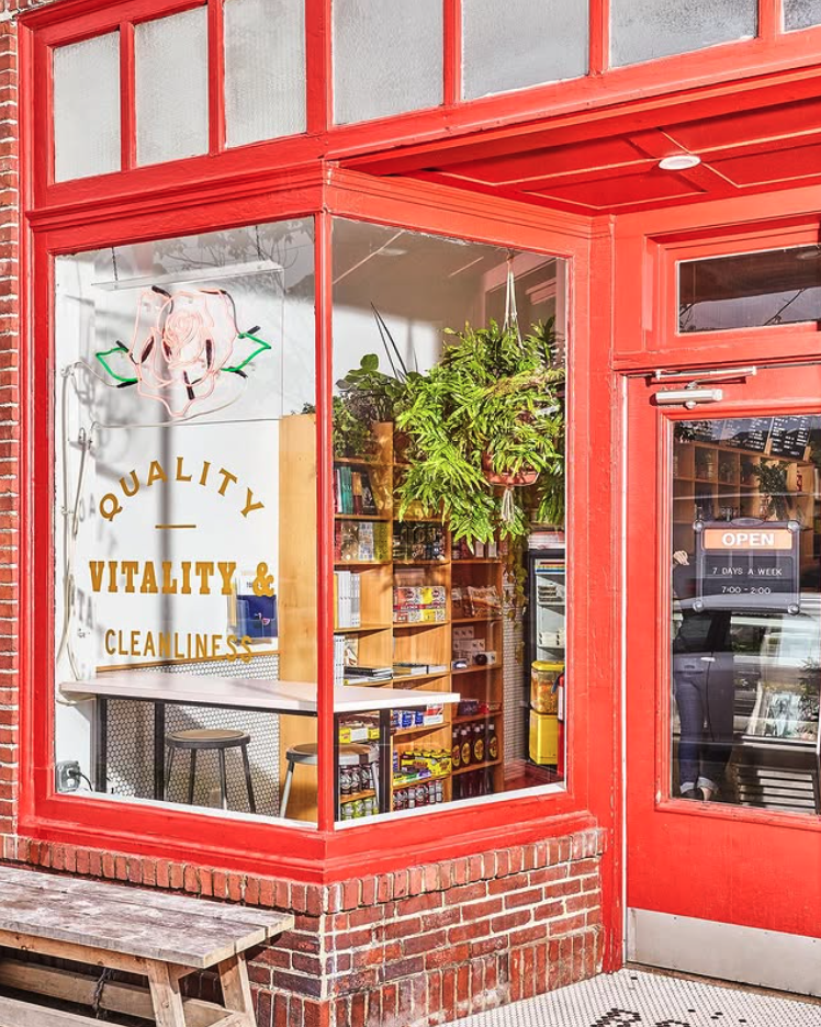 Front view of a store with large glass windows and red framing. Inside, there are shelves with products, a hanging green plant, and a sign that reads 'Quality, Vitality & Cleanliness'. The door has an 'Open' sign and shows a reflection of a person and a car outside.