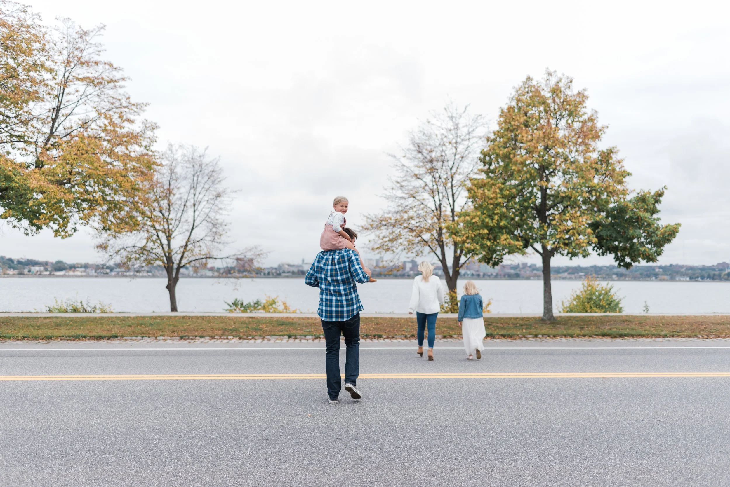 A family walking by a lakeside during fall, with a man carrying a young girl on his shoulders, and two women and a girl walking ahead near the water, trees with fall foliage, overcast sky.