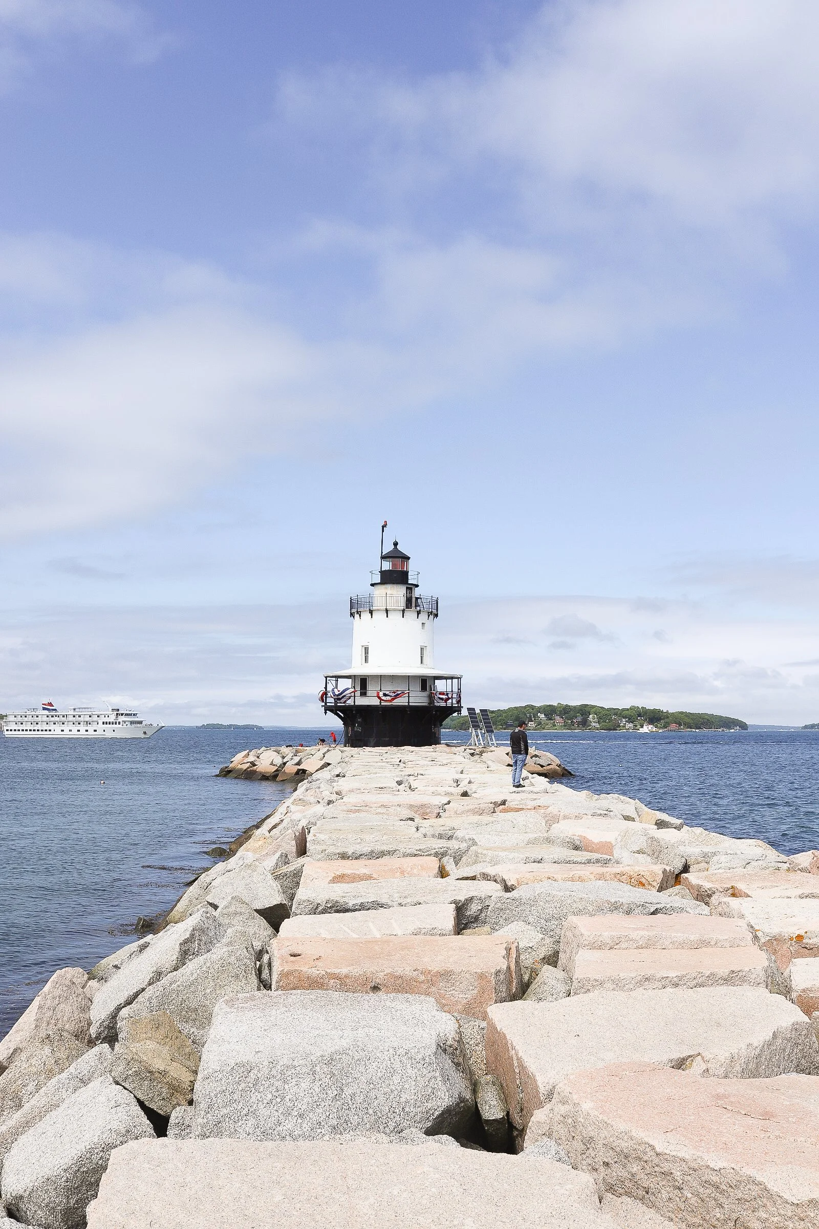 A stone pier leading to a white lighthouse on a sunny day with a blue sky and few clouds, and a boat visible on the water.
