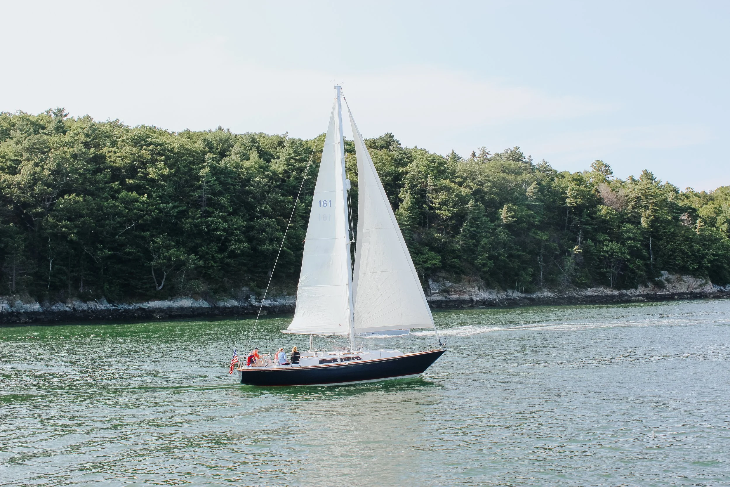 A sailboat with white sails cruising on a body of water with a tree-lined shoreline in the background.