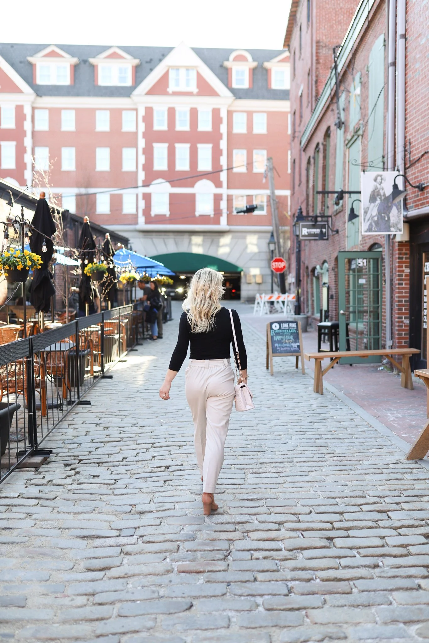 A woman with blonde hair, wearing a black top, beige pants, and carrying a light pink purse, walks down a cobblestone street lined with outdoor dining on the left and brick buildings on the right, with a large brick building with turrets in the background.
