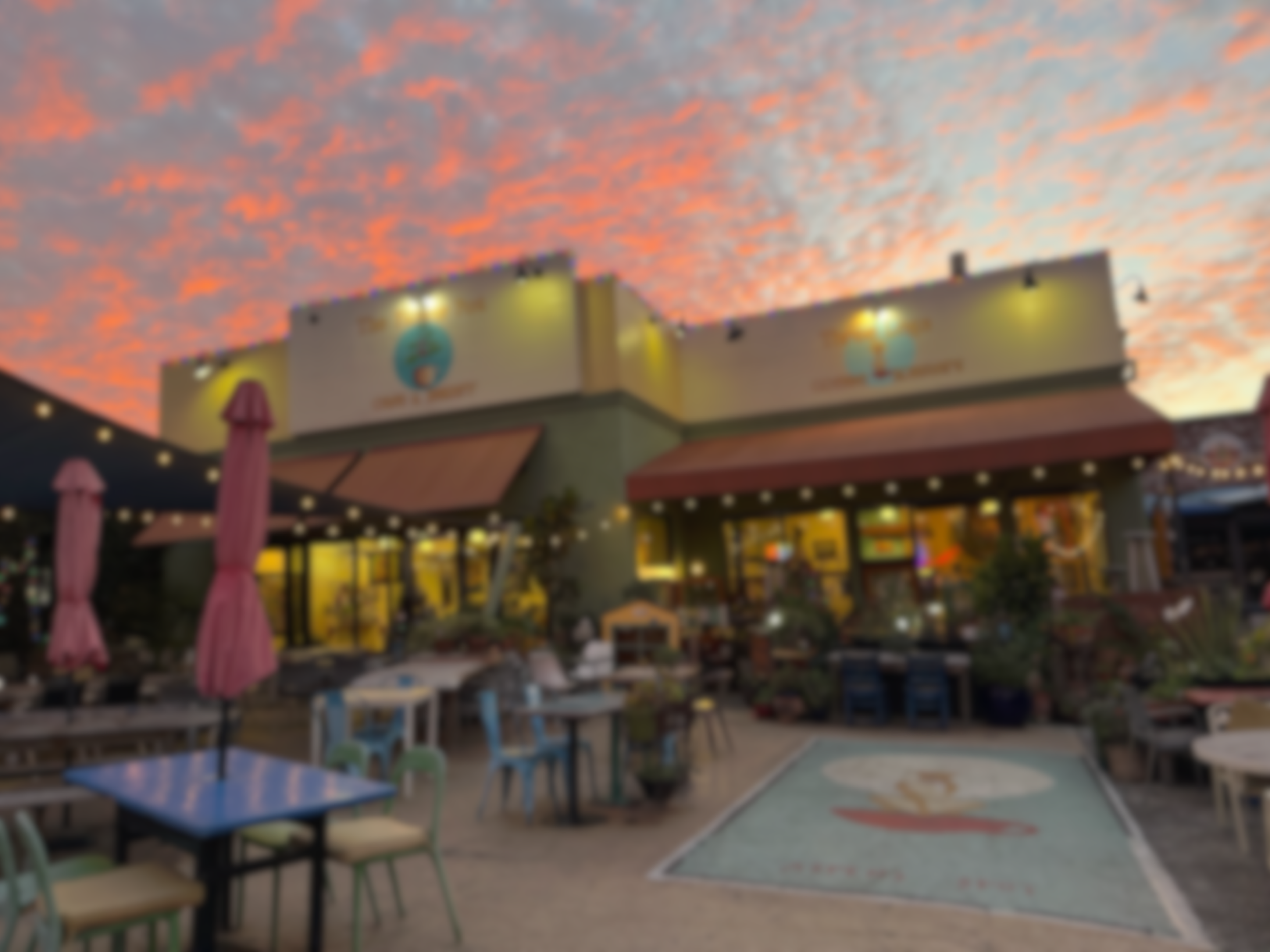 Blurry image of an outdoor restaurant or cafe with tables and umbrellas, a building with a sign, and a colorful sky at sunset.