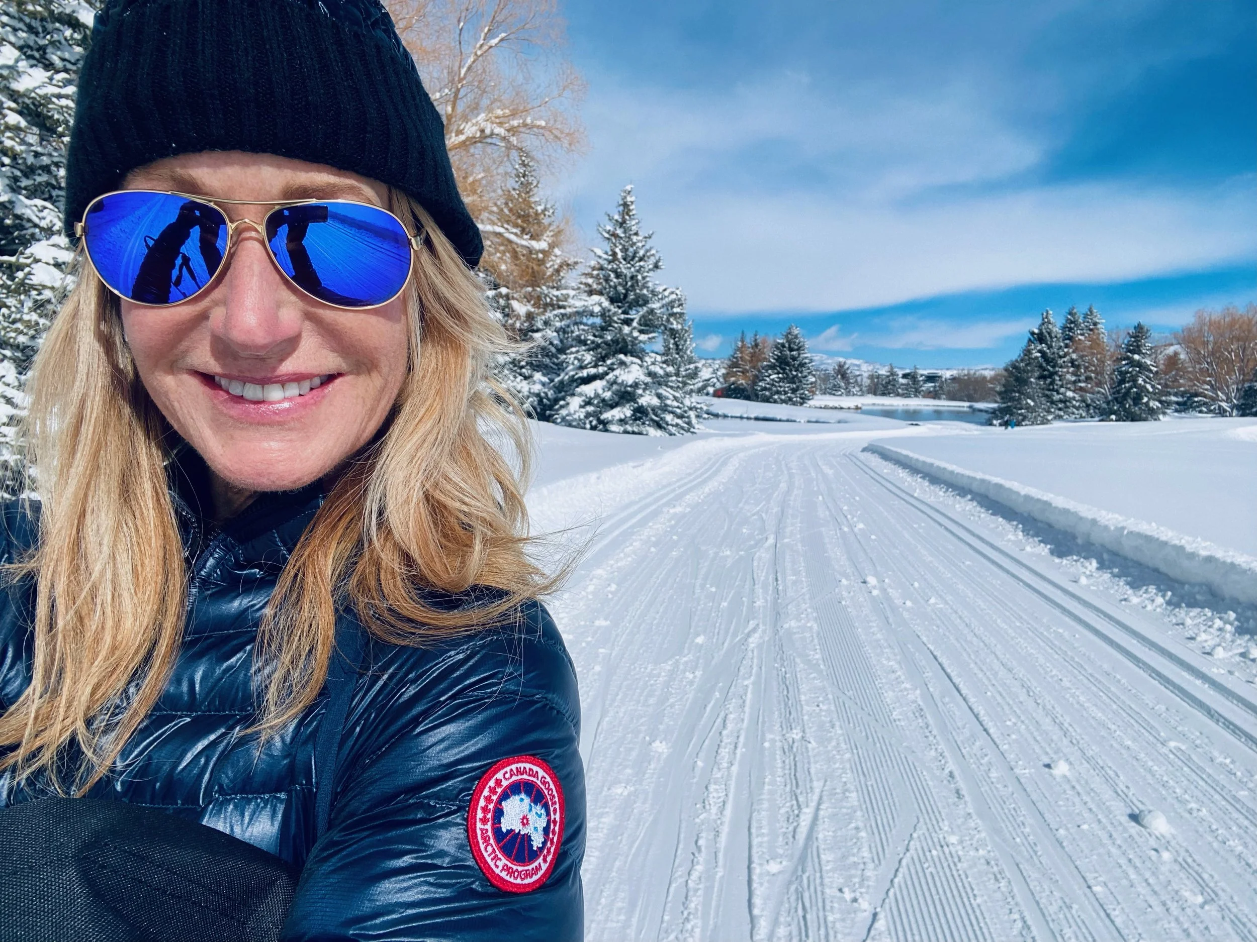 A woman wearing reflective blue sunglasses, a black knit hat, and a navy blue jacket with a Canada Goose logo, smiling in a snowy landscape with snow-covered trees, a frozen path, and a partly cloudy blue sky.