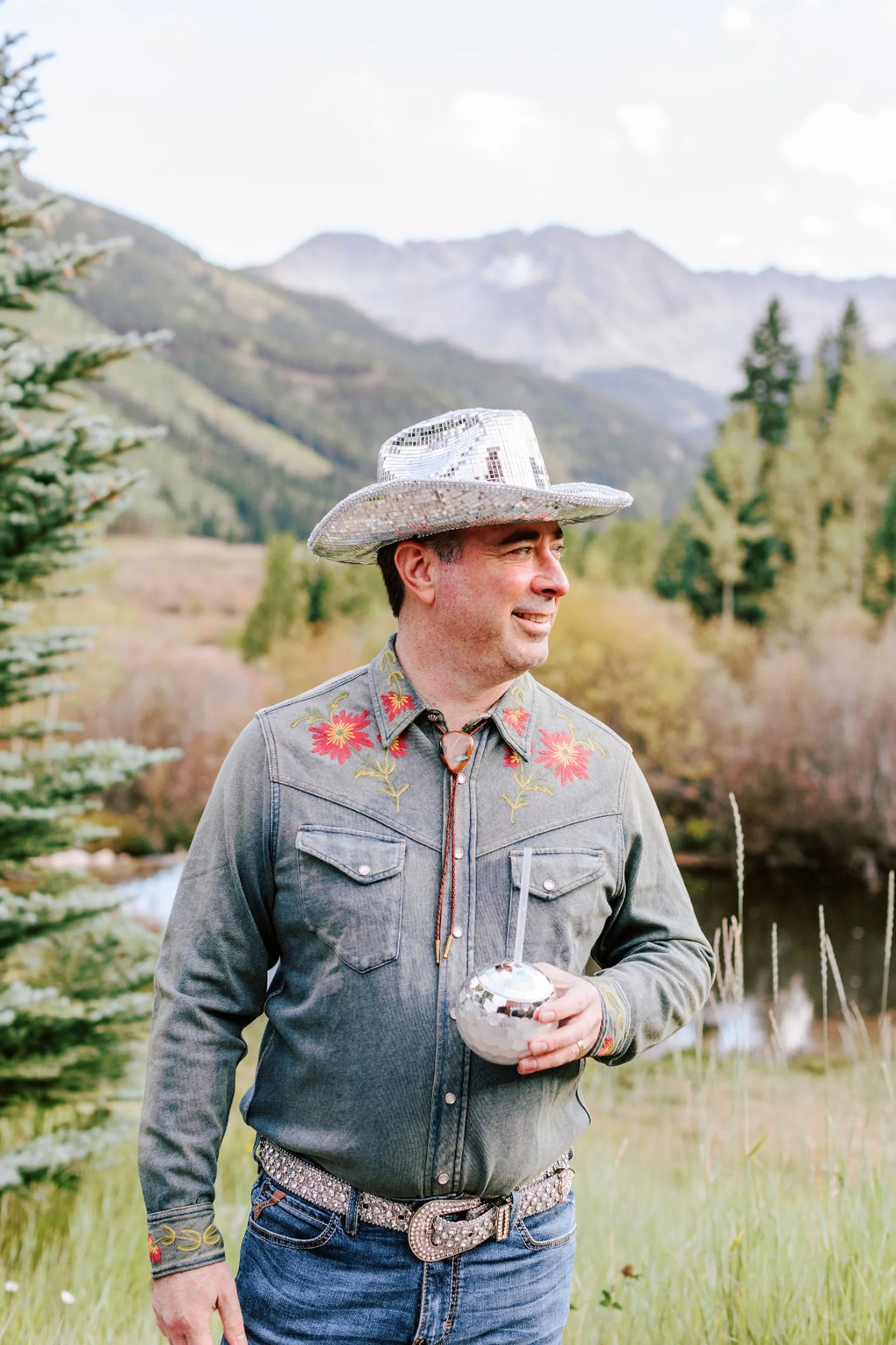 Groom in denim western shirt and silver cowboy hat at the rodeo disco reception at Pine Creek Cookhouse, Elk Mountains in the background, Aspen area wedding