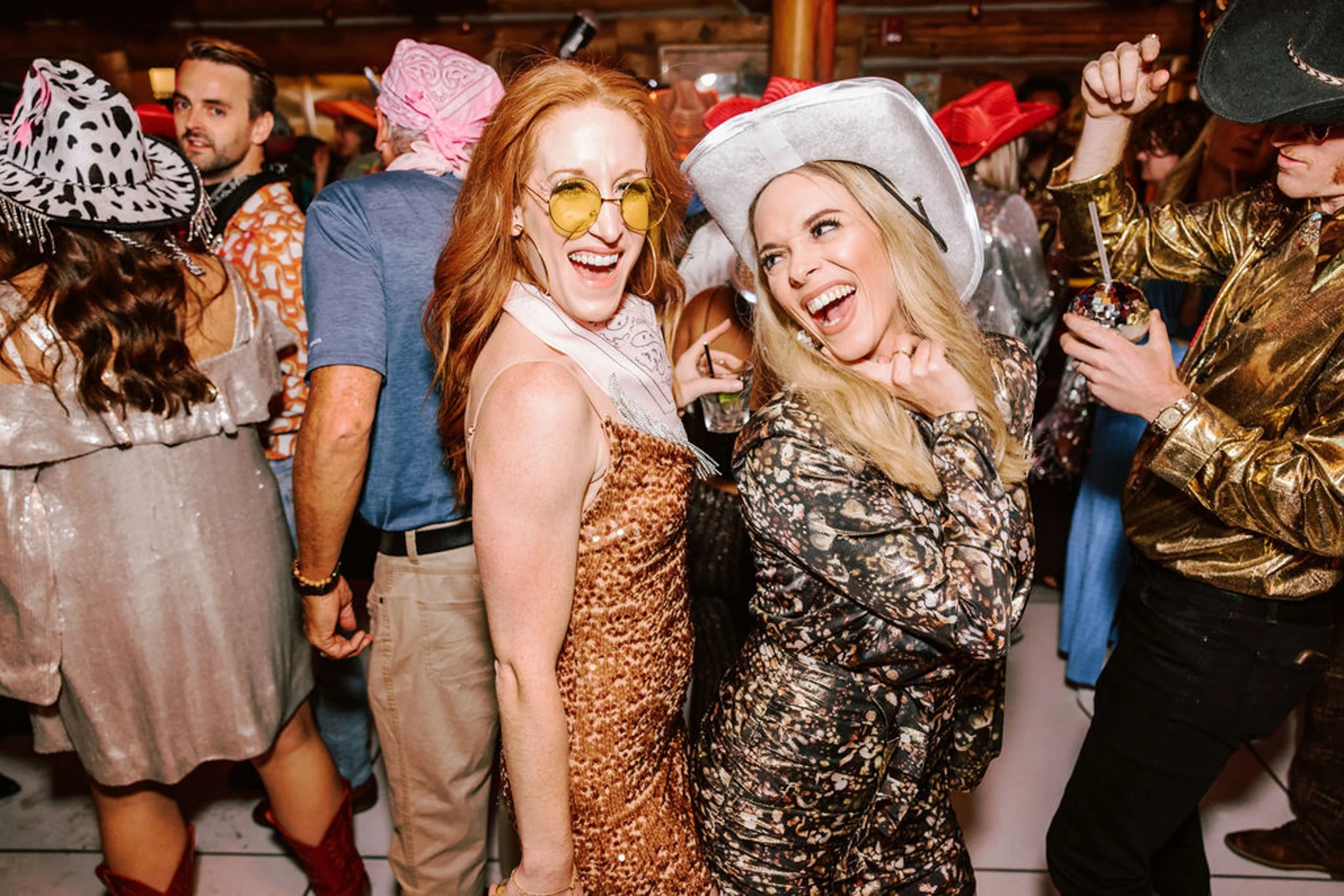 Two women laughing together on the dance floor at Pine Creek Cookhouse wedding reception one in gold sequin dress with yellow sunglasses and one in floral dress with white cowboy hat