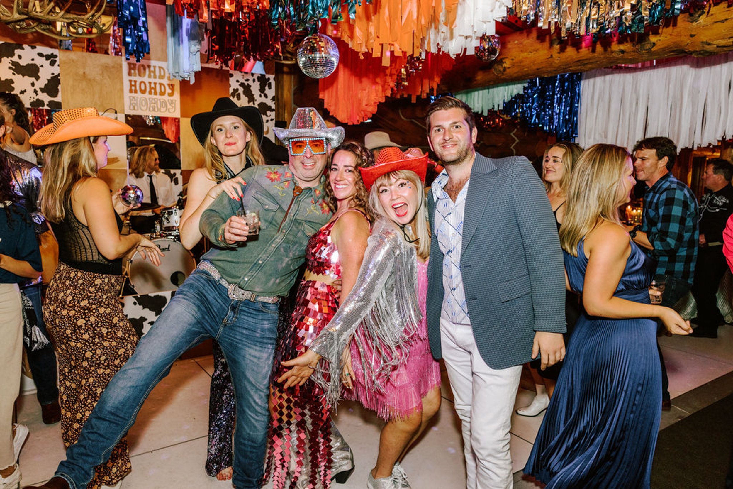 Group of four wedding guests posing in western and sequin outfits in front of Howdy Howdy backdrop at Pine Creek Cookhouse rodeo disco reception with colorful streamers