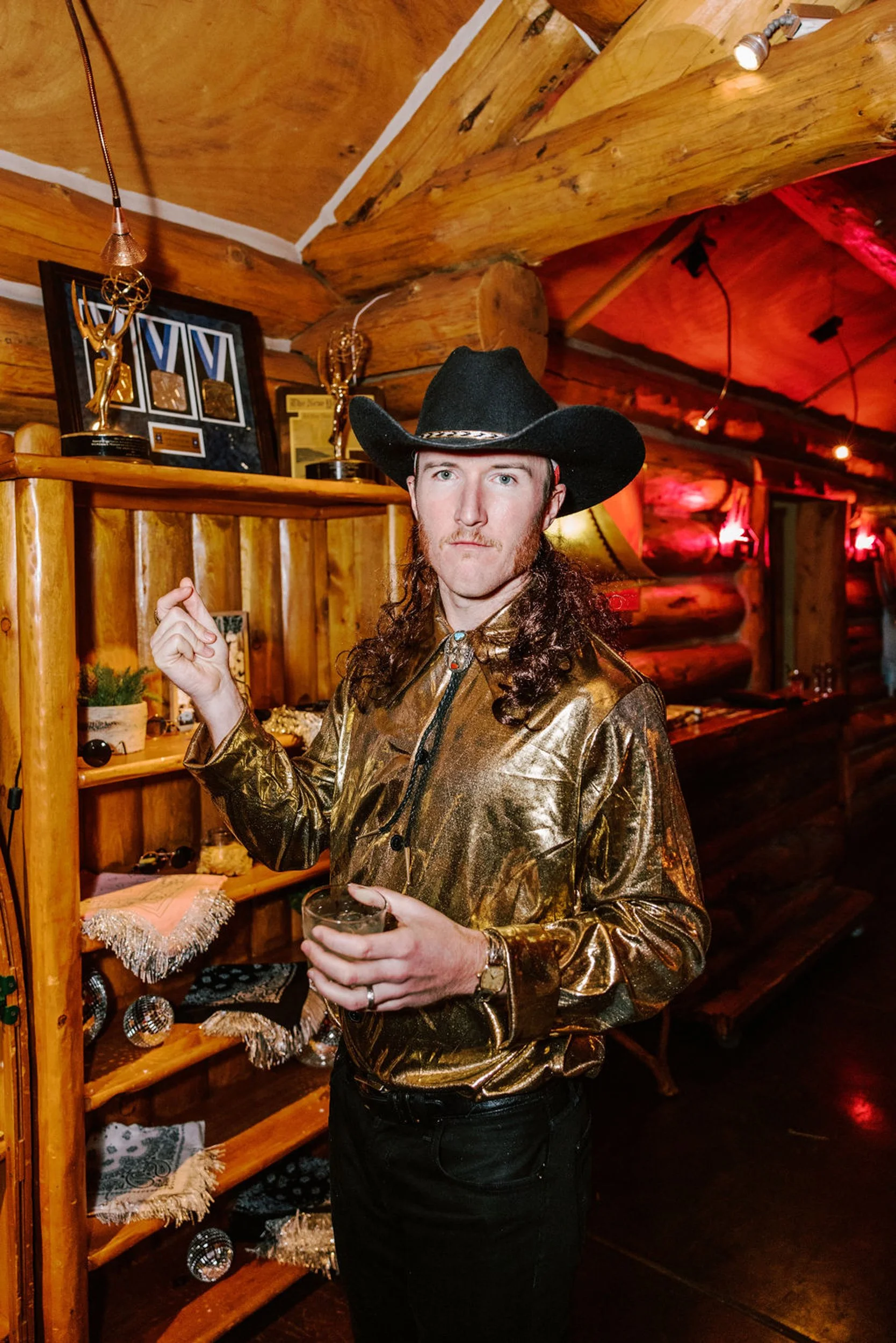 Male guest in gold metallic shirt and black cowboy hat holding a drink inside the rustic log cabin at Pine Creek Cookhouse wedding reception