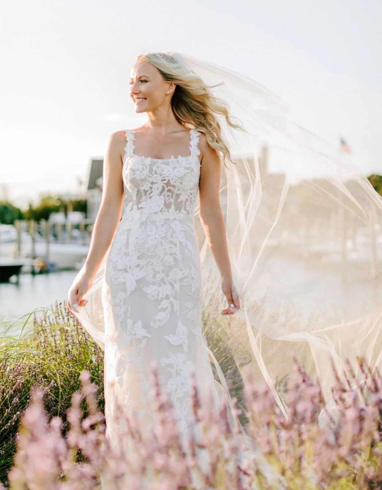 A smiling bride in a white lace wedding dress standing outdoors near water and flowers, with her veil flowing in the breeze.