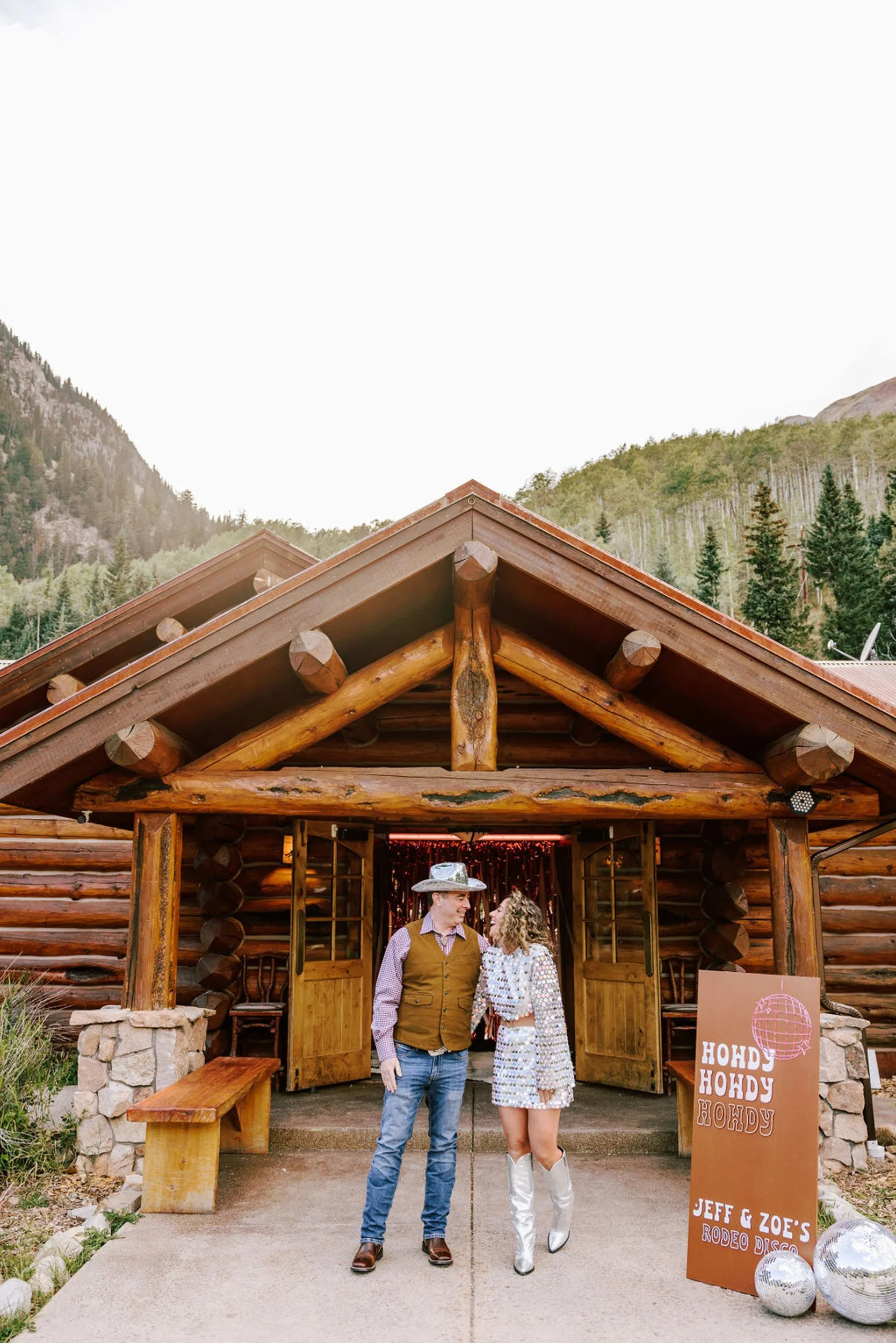 Bride and groom standing under the rustic log entrance of Pine Creek Cookhouse with mountain views behind, Ashcroft Colorado backcountry wedding venue