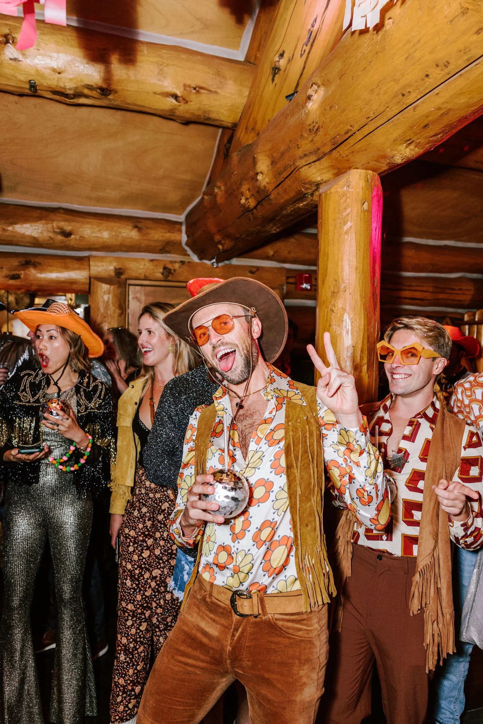 Two male guests in funky western disco outfits with fringe jackets and orange sunglasses celebrating inside Pine Creek Cookhouse log timber reception hall