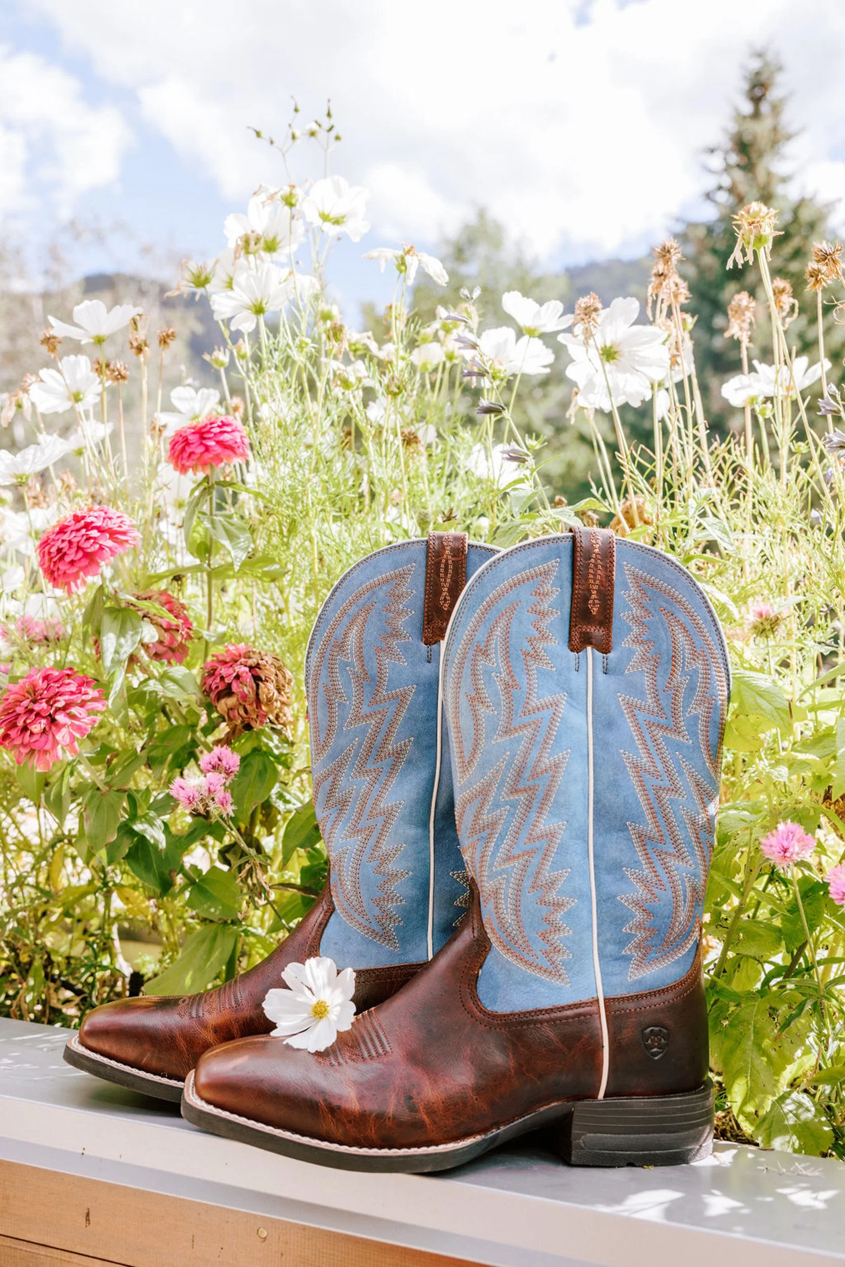 Blue cowboy boots with a white daisy tucked in, displayed in a pink wildflower meadow at Pine Creek Cookhouse, rodeo disco wedding styling detail