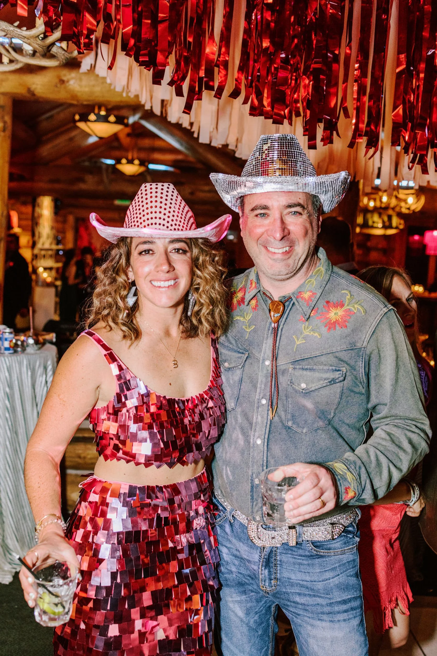 Couple in matching sequin cowboy hats posing together at Pine Creek Cookhouse wedding reception woman in red sequin outfit and man in embroidered western denim shirt