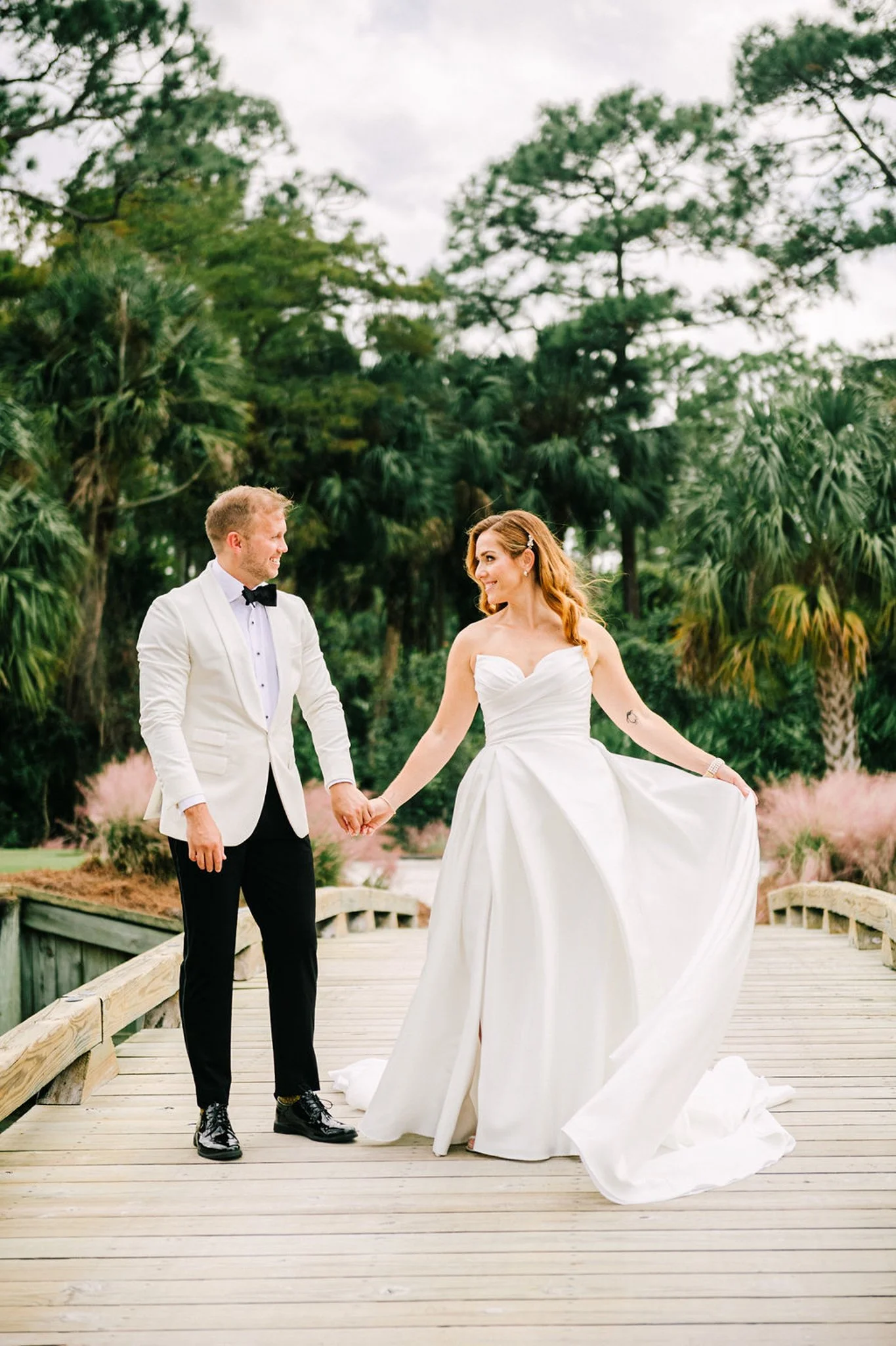 A bride and groom on their wedding day. the couple is holding hands on a wooden bridge in a lush, green outdoor setting, with the bride wearing a strapless white wedding gown and the groom in a white tuxedo jacket and black pants.