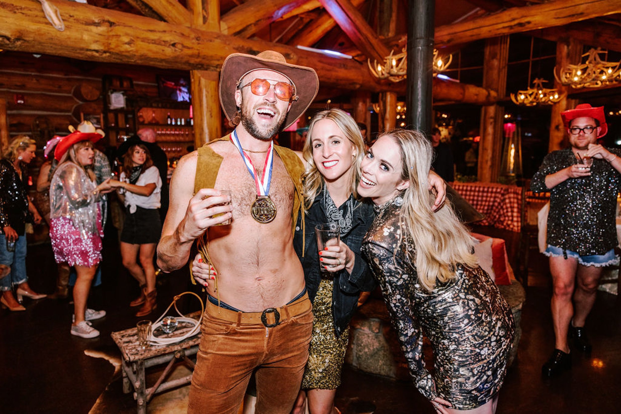 Playful wedding guest in cowboy hat and vest with a medal posing with two women in sparkly outfits inside Pine Creek Cookhouse log cabin reception