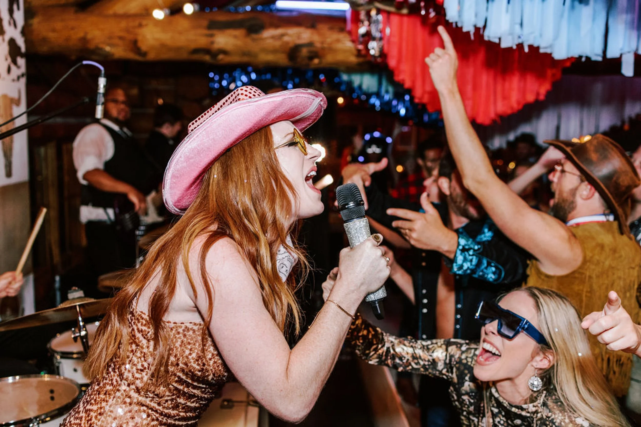 Female wedding guest in pink cowboy hat and gold sequin dress singing into a microphone with joyful guests and live band at Pine Creek Cookhouse wedding reception
