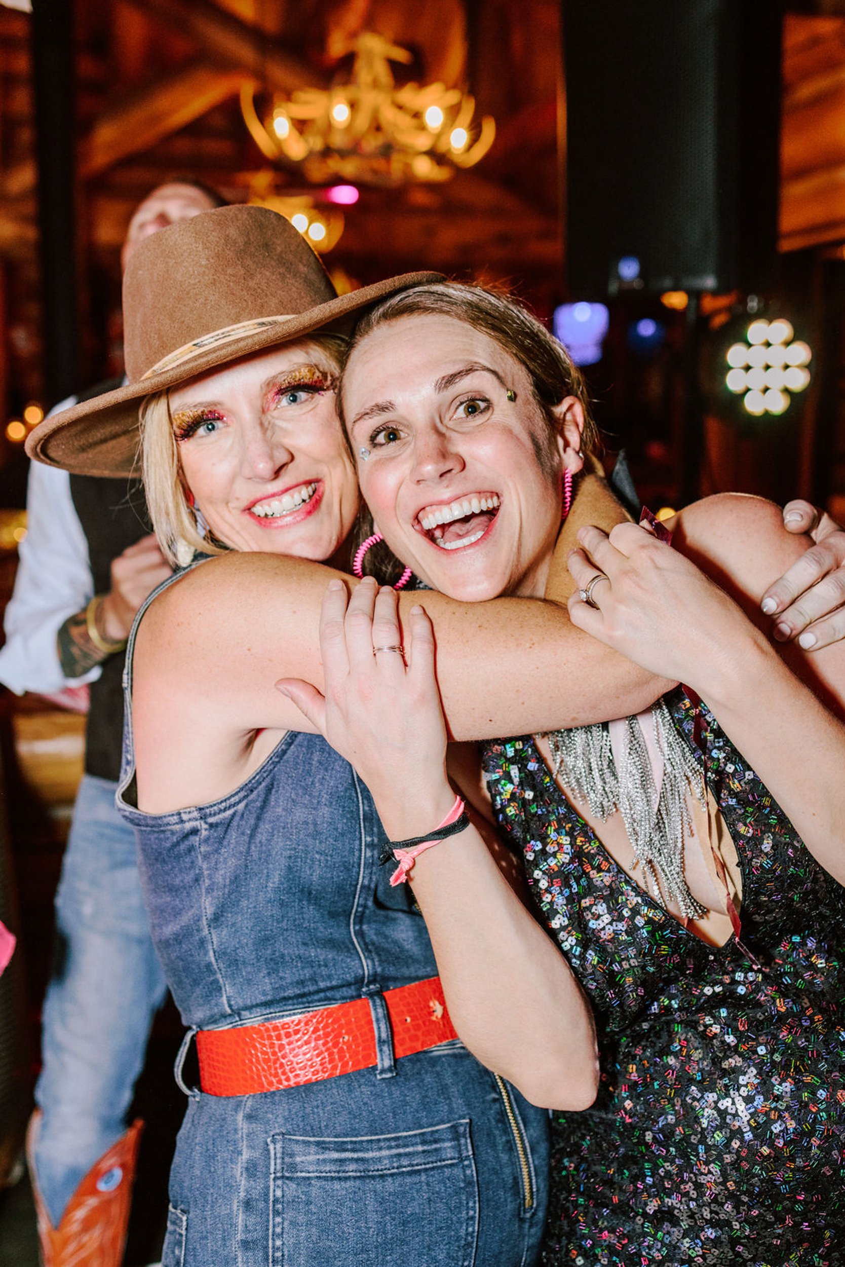 Two female wedding guests laughing and hugging at Pine Creek Cookhouse reception one in denim dress with tan cowboy hat and one in dark sparkly dress