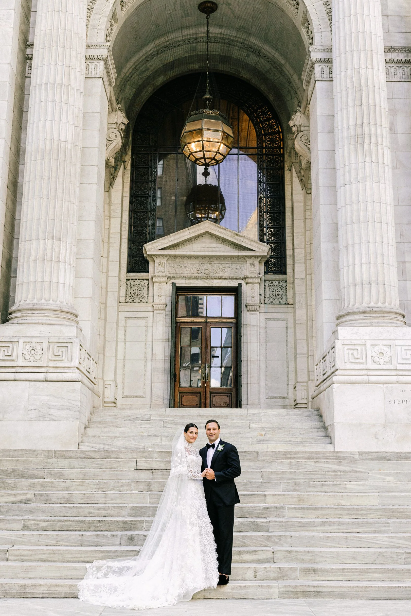 Bride and groom standing on the exterior steps of the New York Public Library with the ornate columns and bronze doors behind them