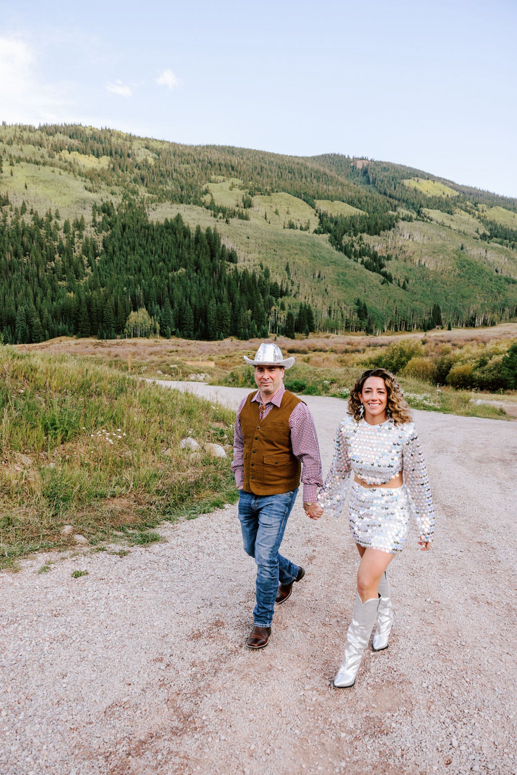 Newlyweds in reception outfits walking a mountain road with the Elk Mountains and Castle Creek Valley behind them, Pine Creek Cookhouse Aspen wedding