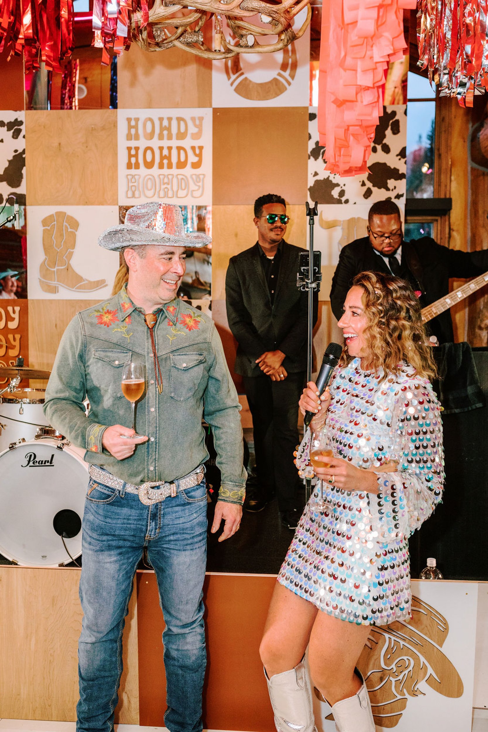 Couple laughing together in front of Howdy Howdy Howdy photo backdrop with live band behind them at Pine Creek Cookhouse rodeo disco wedding reception