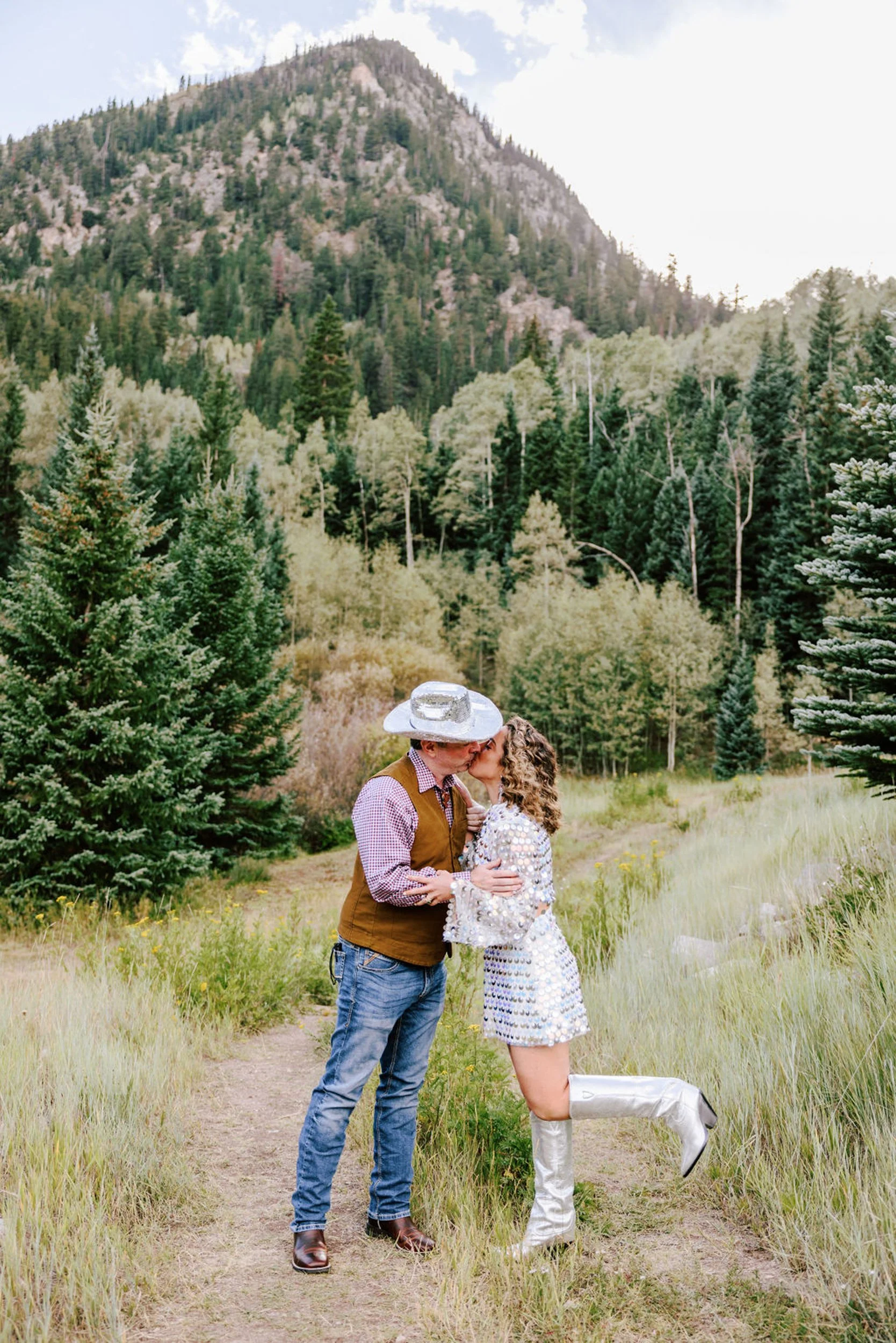 Newlyweds in reception outfits walking a pine-lined path at Pine Creek Cookhouse, groom in cowboy hat, Elk Mountains behind them, Aspen area wedding photography